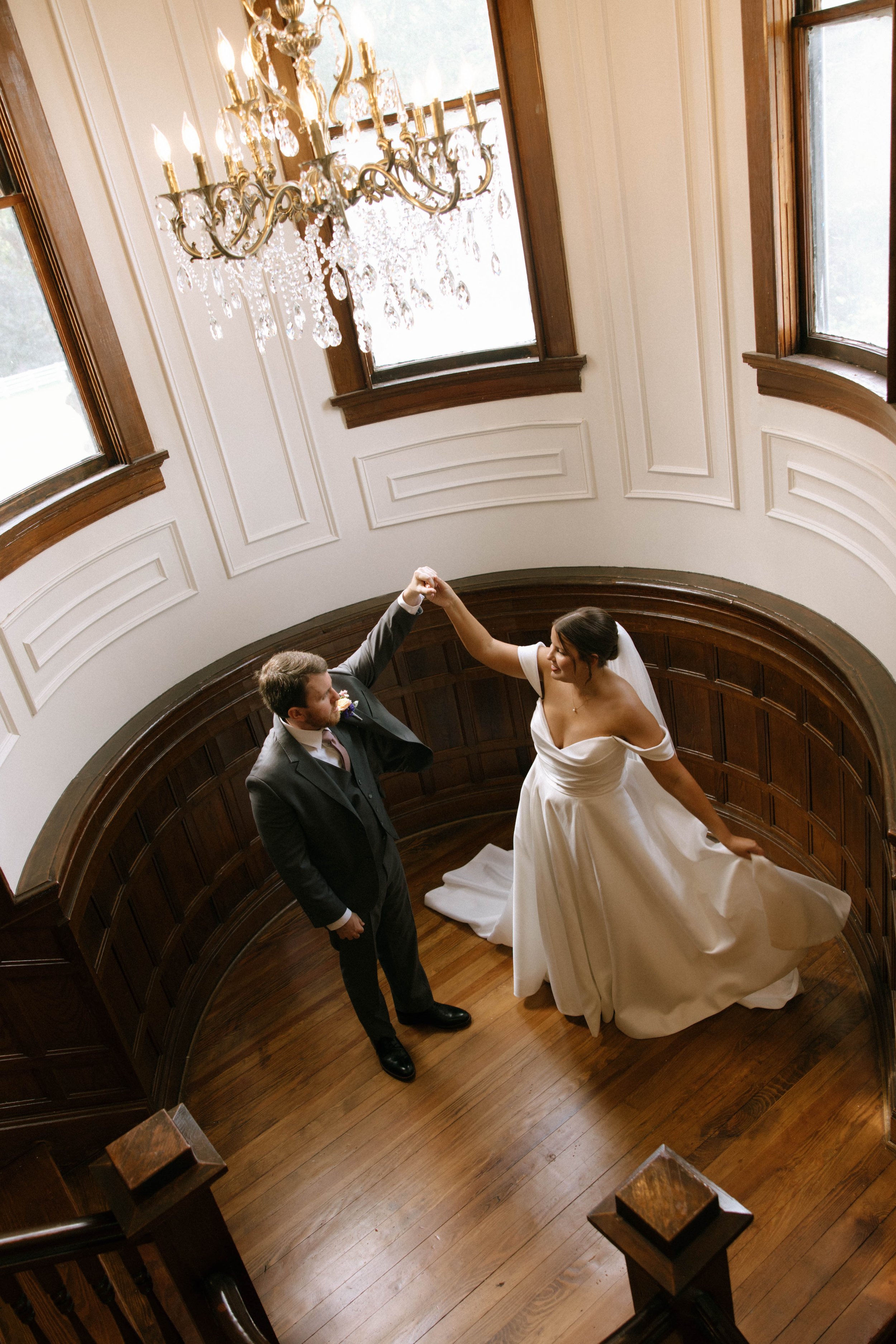 A bride and groom dancing on a wooden staircase with carved wood paneling, chandelier, and large windows.