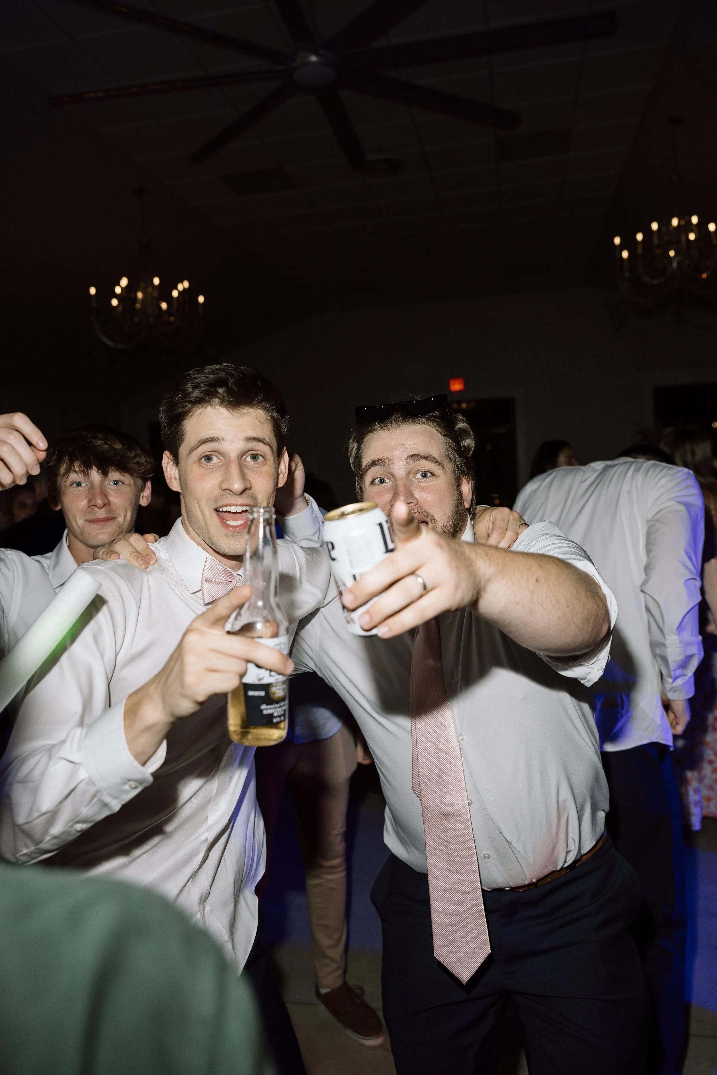 Two men smiling and holding drinks at a lively party, with other guests in the background.