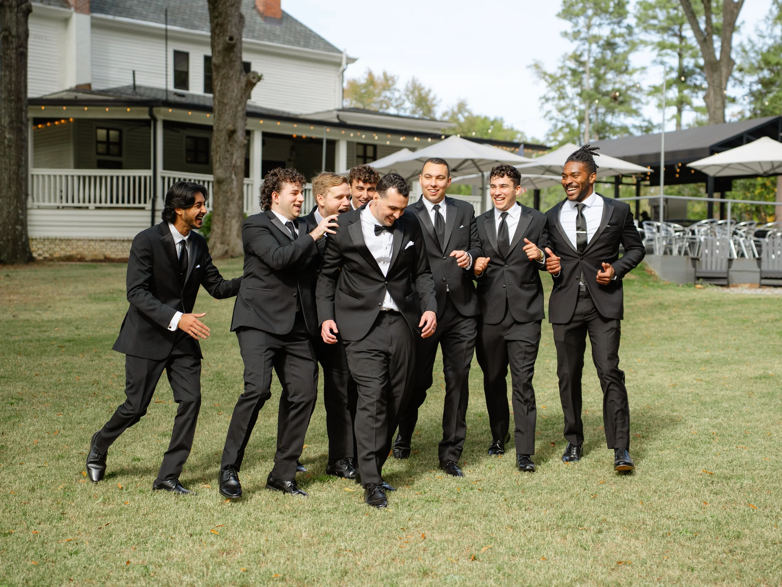 Group of men in black suits walking on grass, laughing and celebrating outdoors at a formal event.