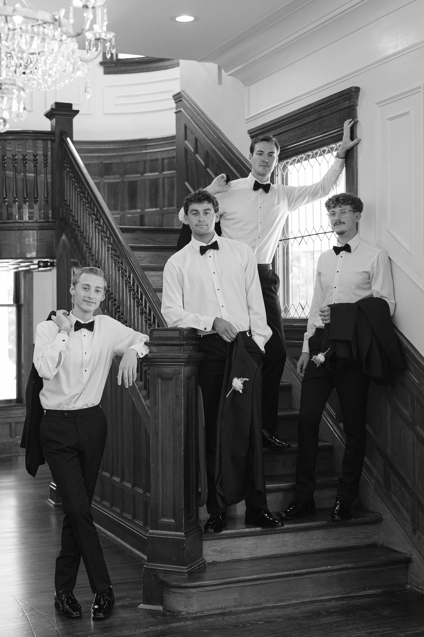 Five young men in tuxedos with bow ties, posing on a wooden staircase inside a fancy building with a chandelier and large window.