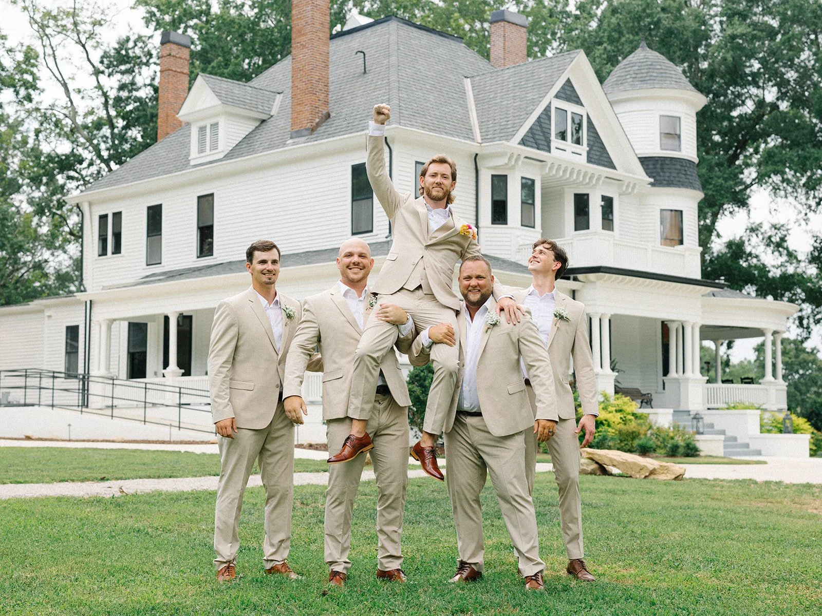 Group of five men in beige suits, one being lifted on shoulders, in front of a large white Victorian style house with a wraparound porch and turret, green lawn, trees, and cloudy sky.