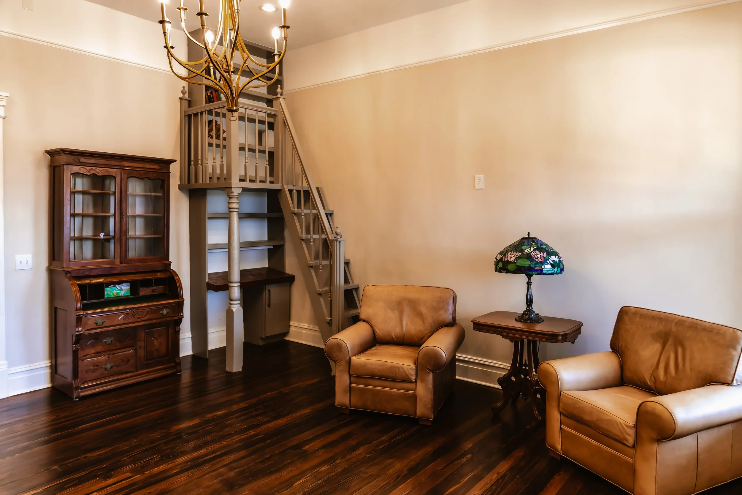 Living room with two tan leather armchairs, a wooden side table with a stained glass lamp, a wooden china cabinet, and a staircase with a small dining area underneath.