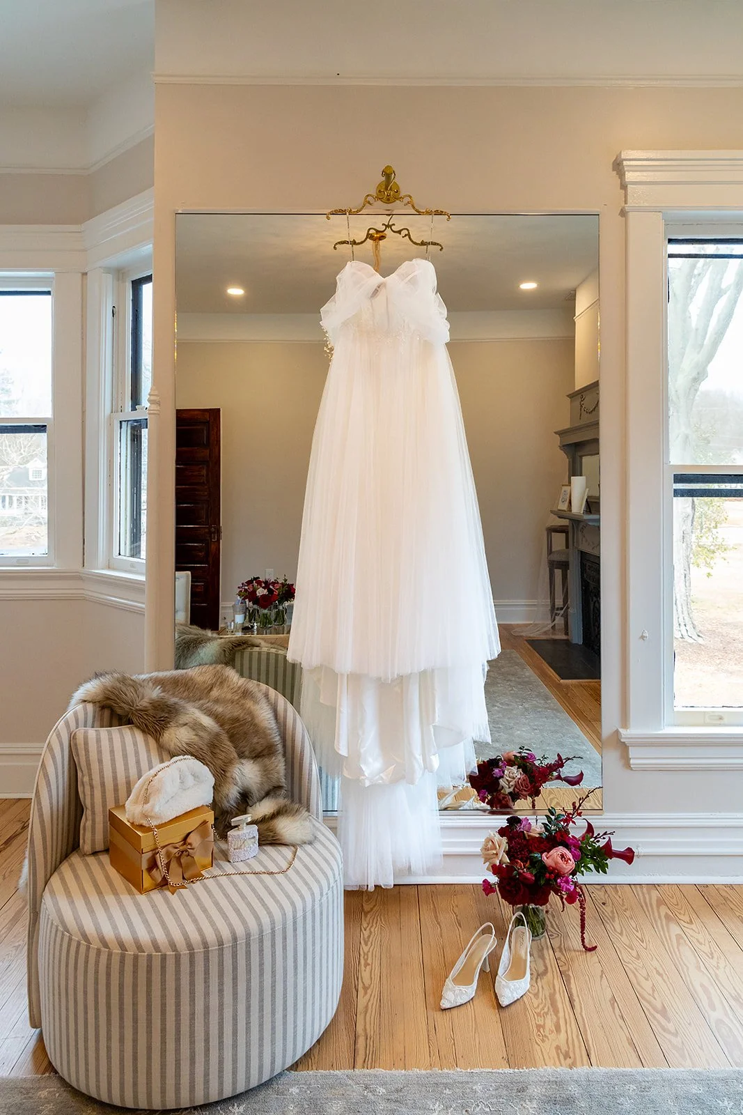 A white wedding dress hanging in front of a large mirror, with a pair of white shoes and a floral bouquet on the floor, a chair with a fur throw and a gift box on it, and a window showing trees outside.