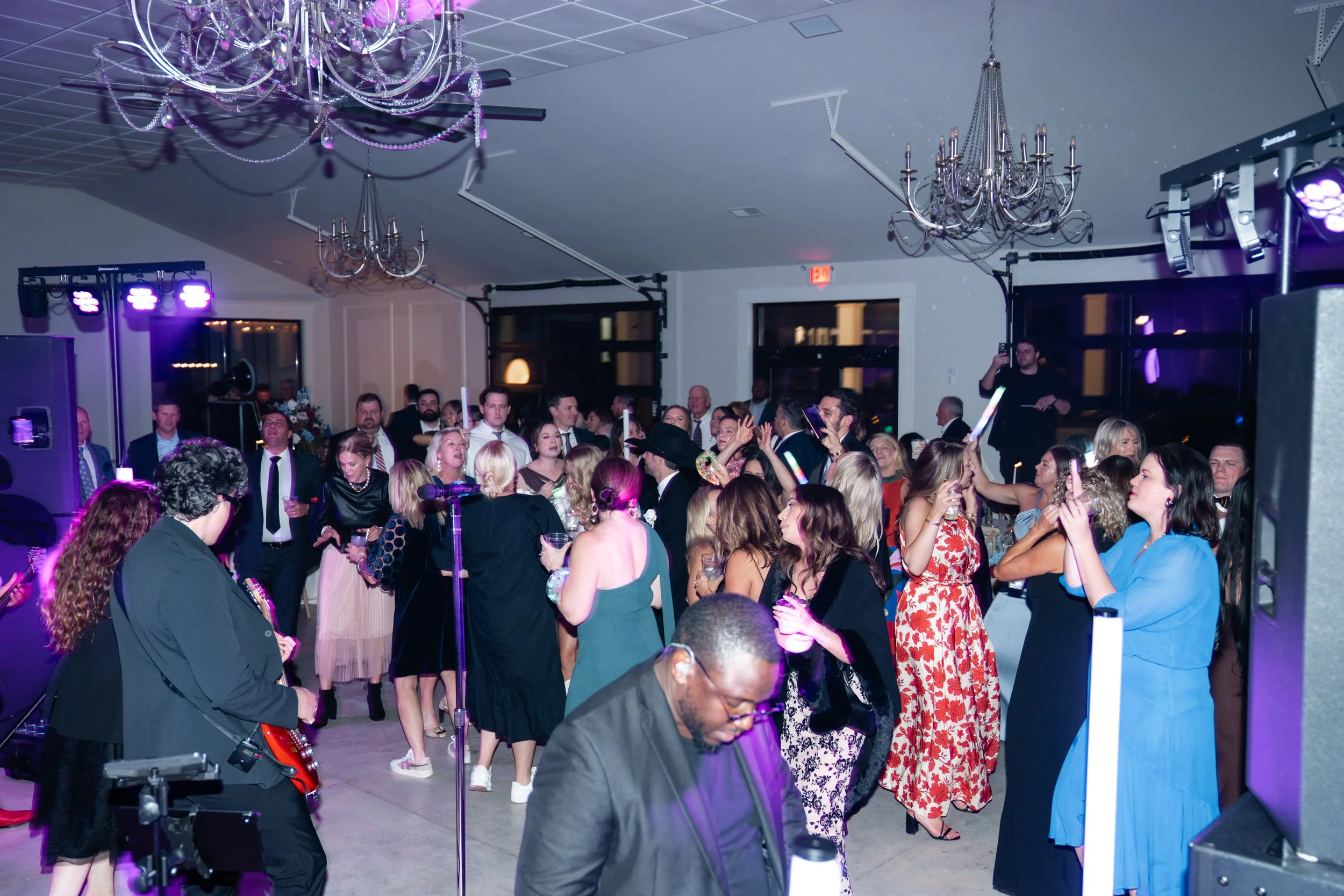 People dancing and socializing at a party or celebration in an indoor venue with chandeliers, purple lighting, and DJ equipment.