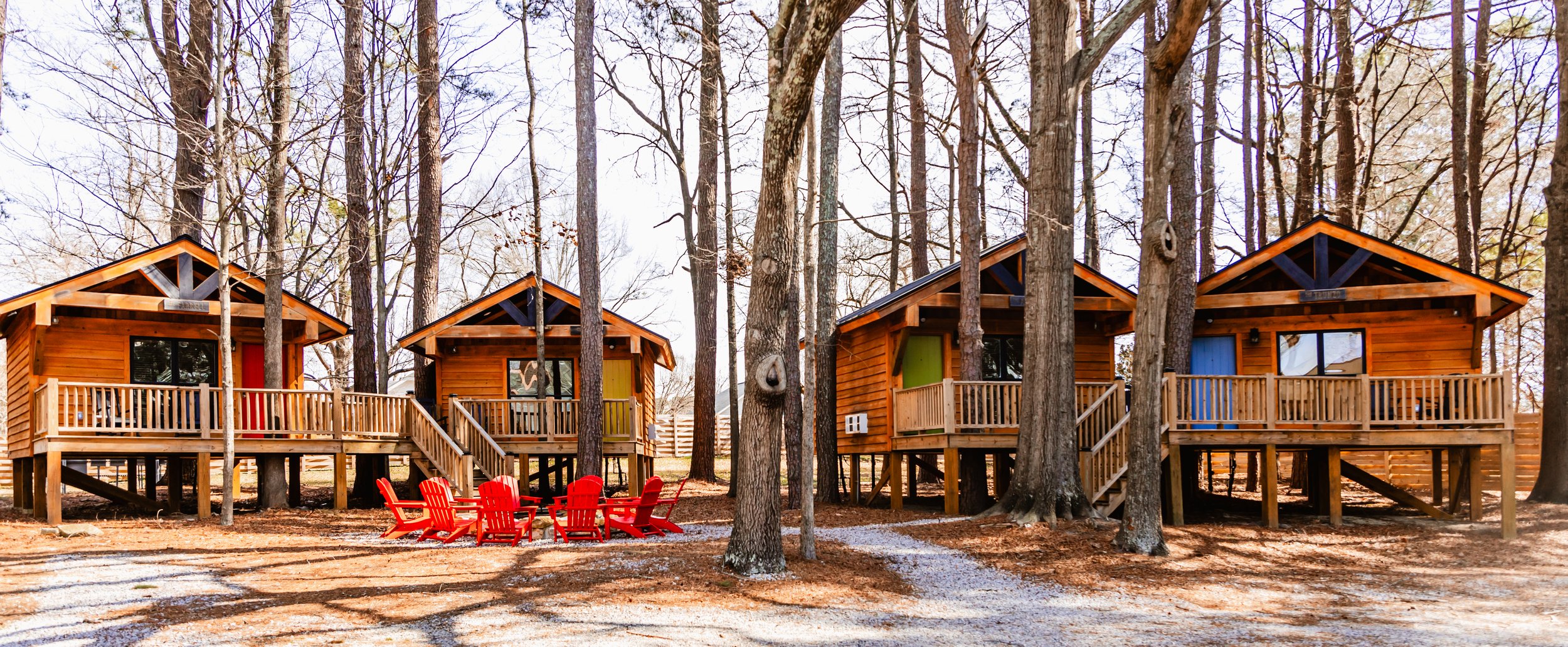 Four wooden cabins in a forest with tall trees, each with a small roofed porch and stairs leading to the ground, and red Adirondack chairs on the ground in front of the cabins.