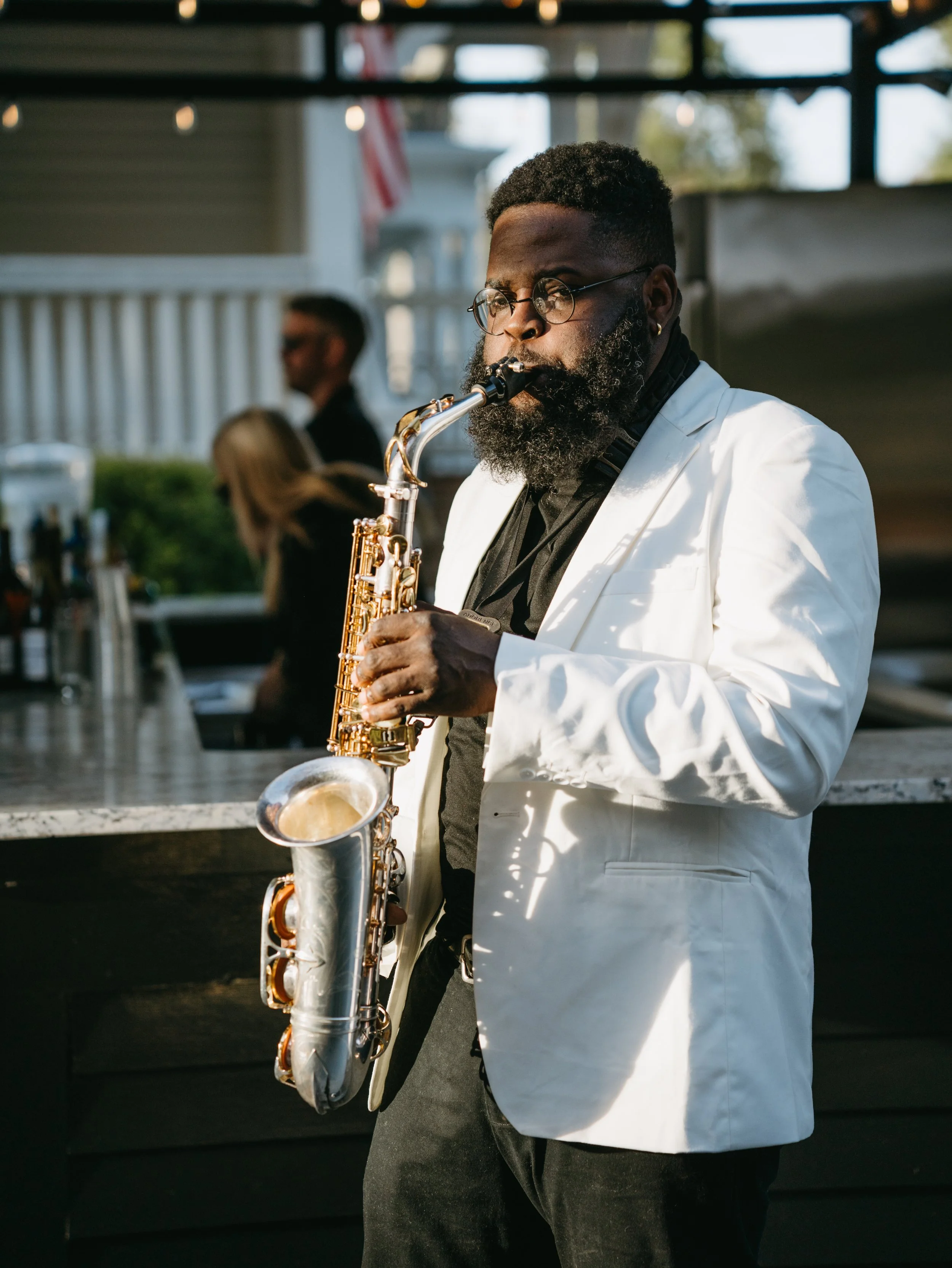 A man with glasses and a beard, dressed in a white blazer, playing saxophone outdoors during daytime.