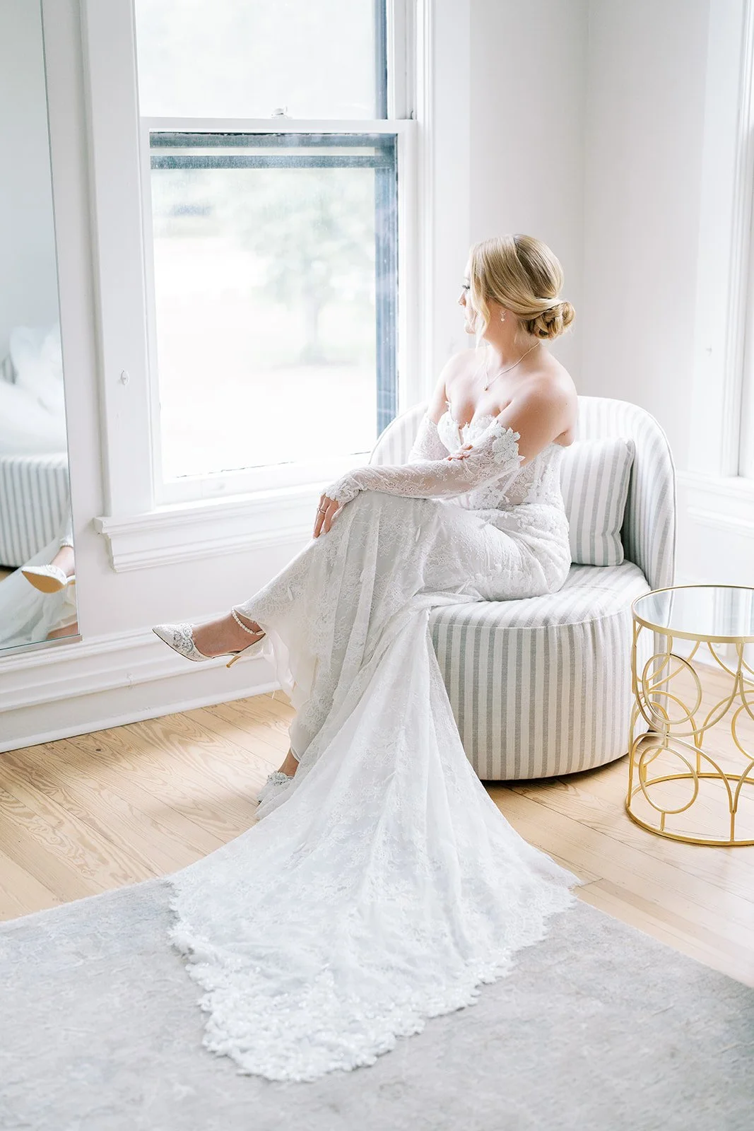 A bride in a white lace wedding dress sitting on a striped chair by a large window, looking outside, with her legs crossed and wearing elegant heels and jewelry.