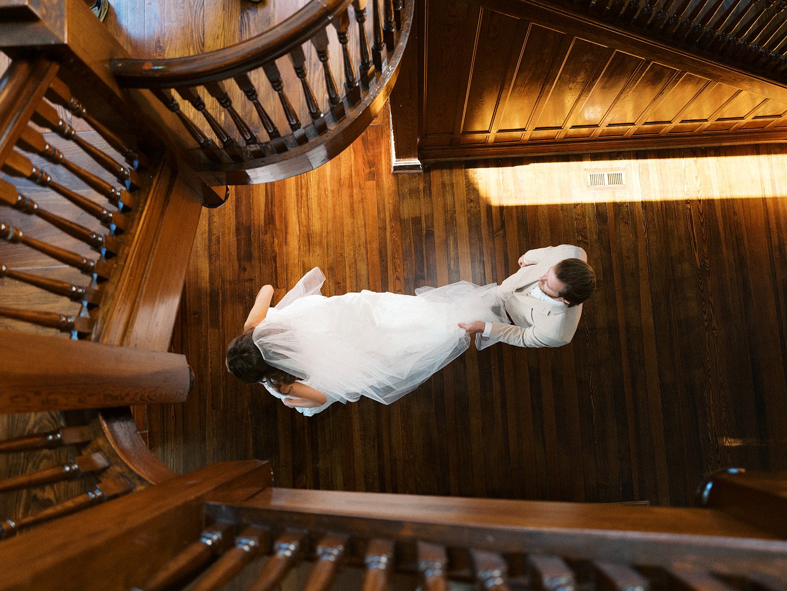 A bride and groom standing on a wooden floor, viewed from above, with the bride in a white wedding dress and the groom in a light-colored suit.
