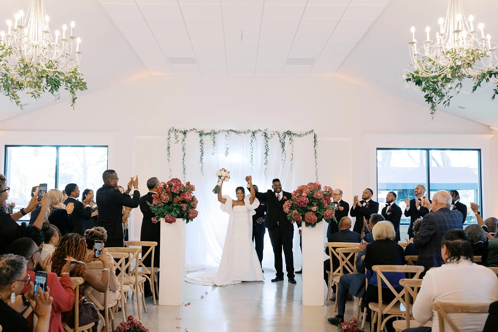 A wedding ceremony with the bride and groom celebrating on stage, surrounded by guests clapping and taking pictures in a bright, decorated venue with large windows and chandeliers.