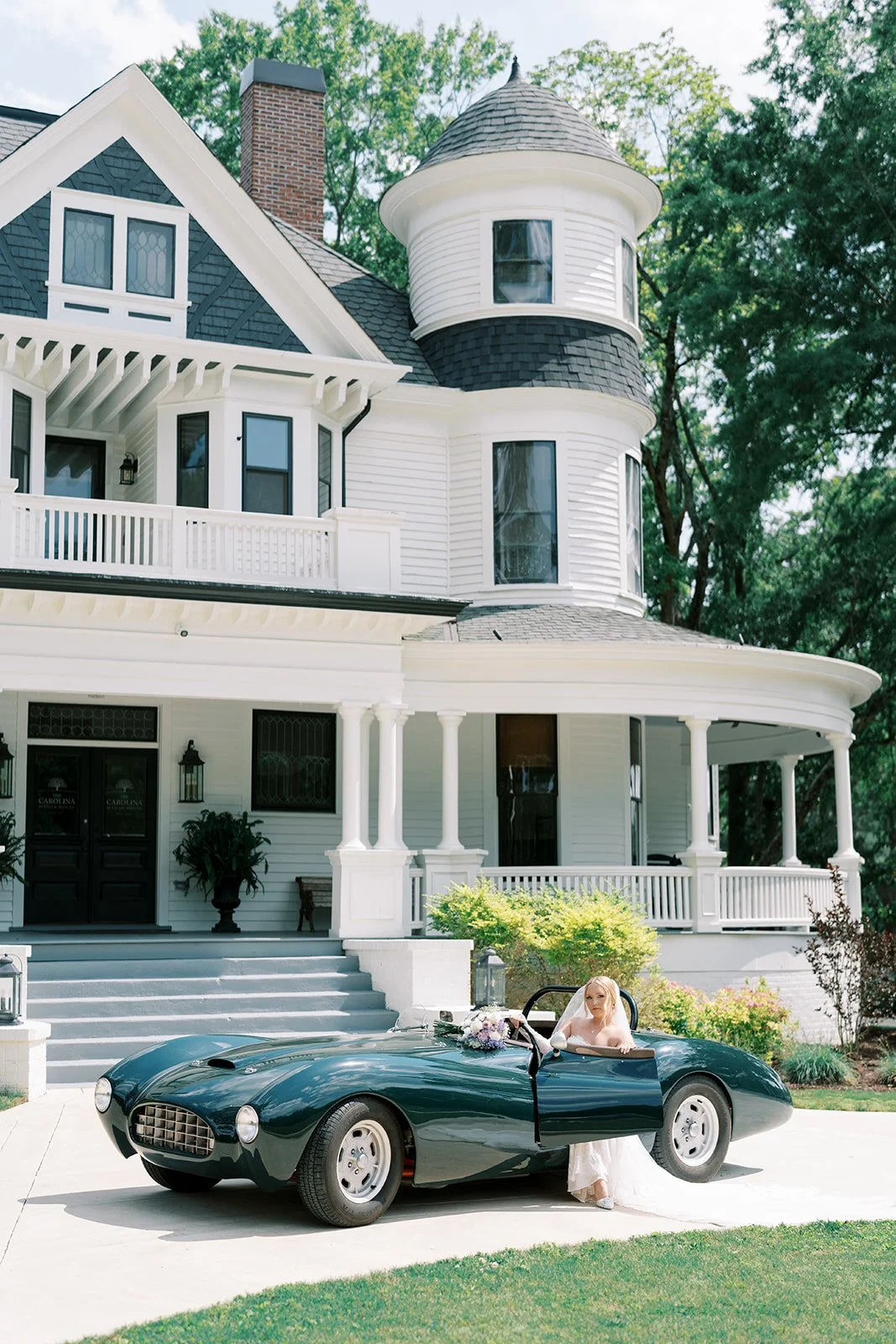 A bride sitting in a vintage black sports car, parked in front of a large, white Victorian-style house with a turret.