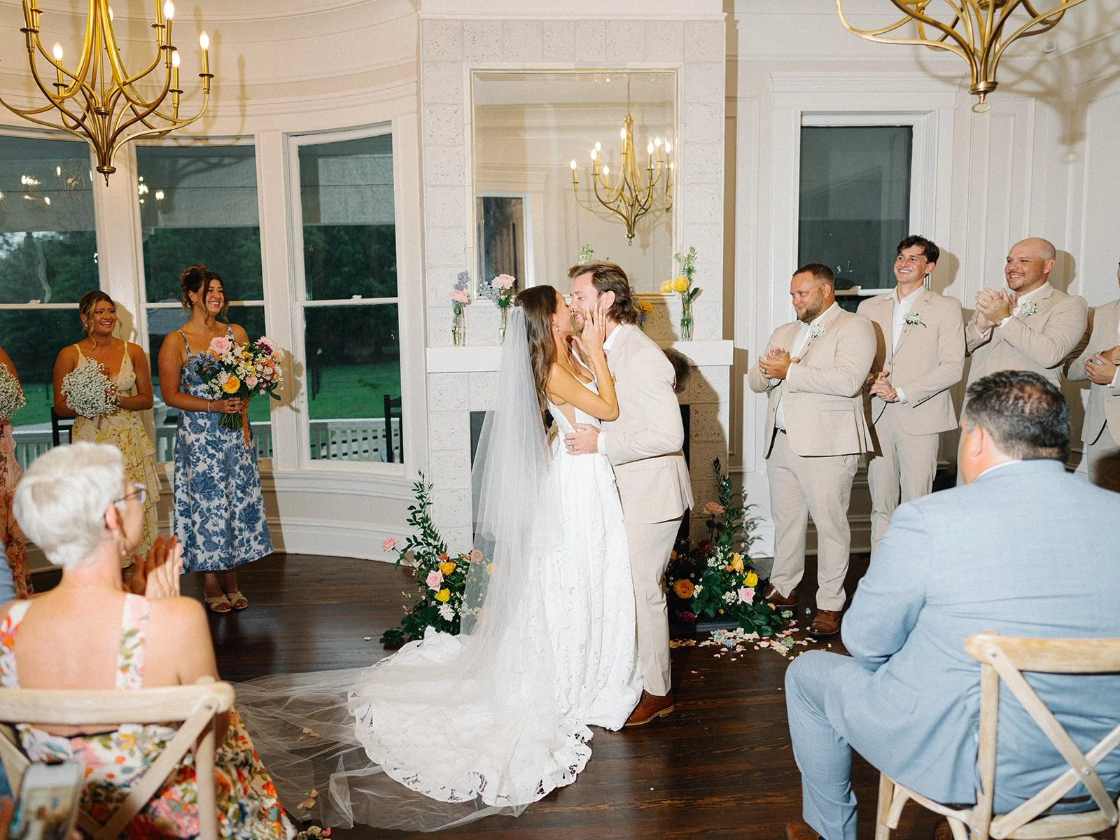 A wedding ceremony with a bride and groom sharing a kiss in front of guests, including bridesmaids and groomsmen, inside a bright, elegantly decorated room with large windows, floral arrangements, and chandeliers.
