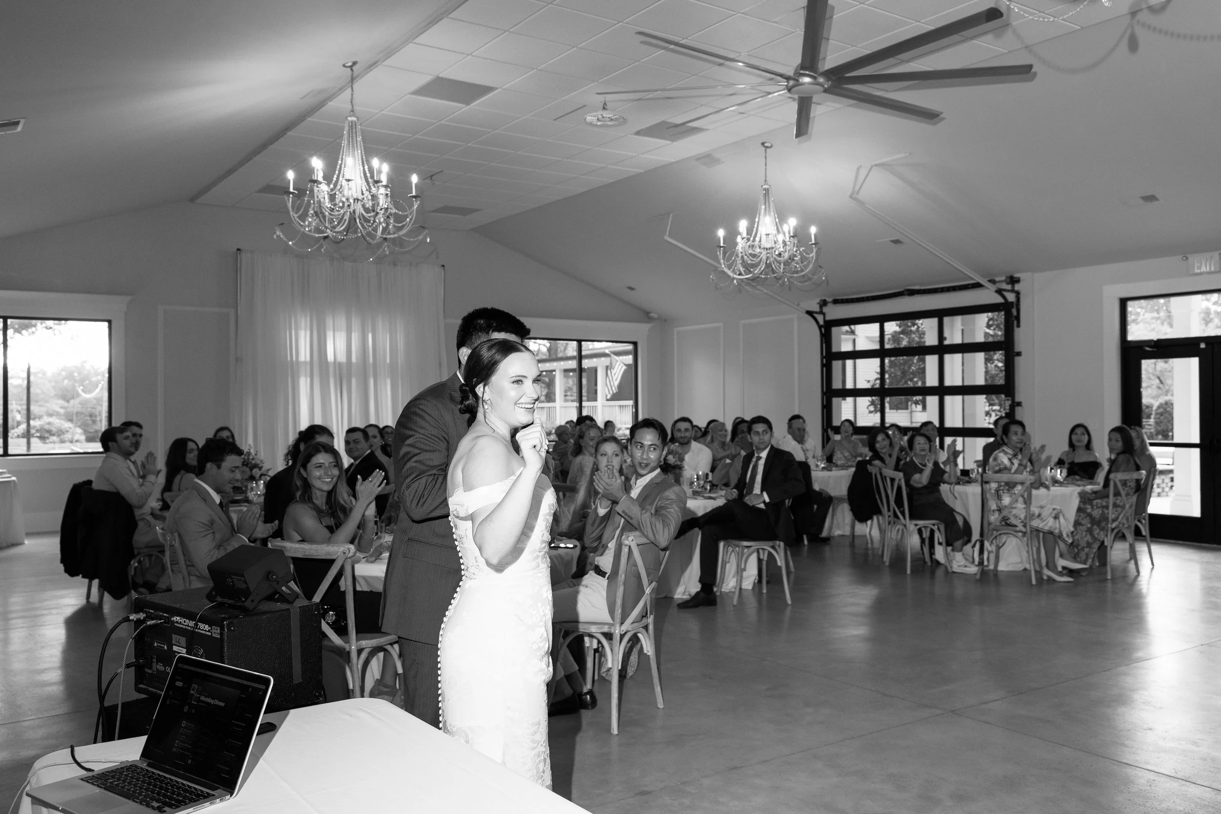 A bride and groom standing together at their wedding reception, smiling and enjoying their moment, with guests seated at round tables clapping and smiling in the background, under chandeliers in a decorated banquet hall.
