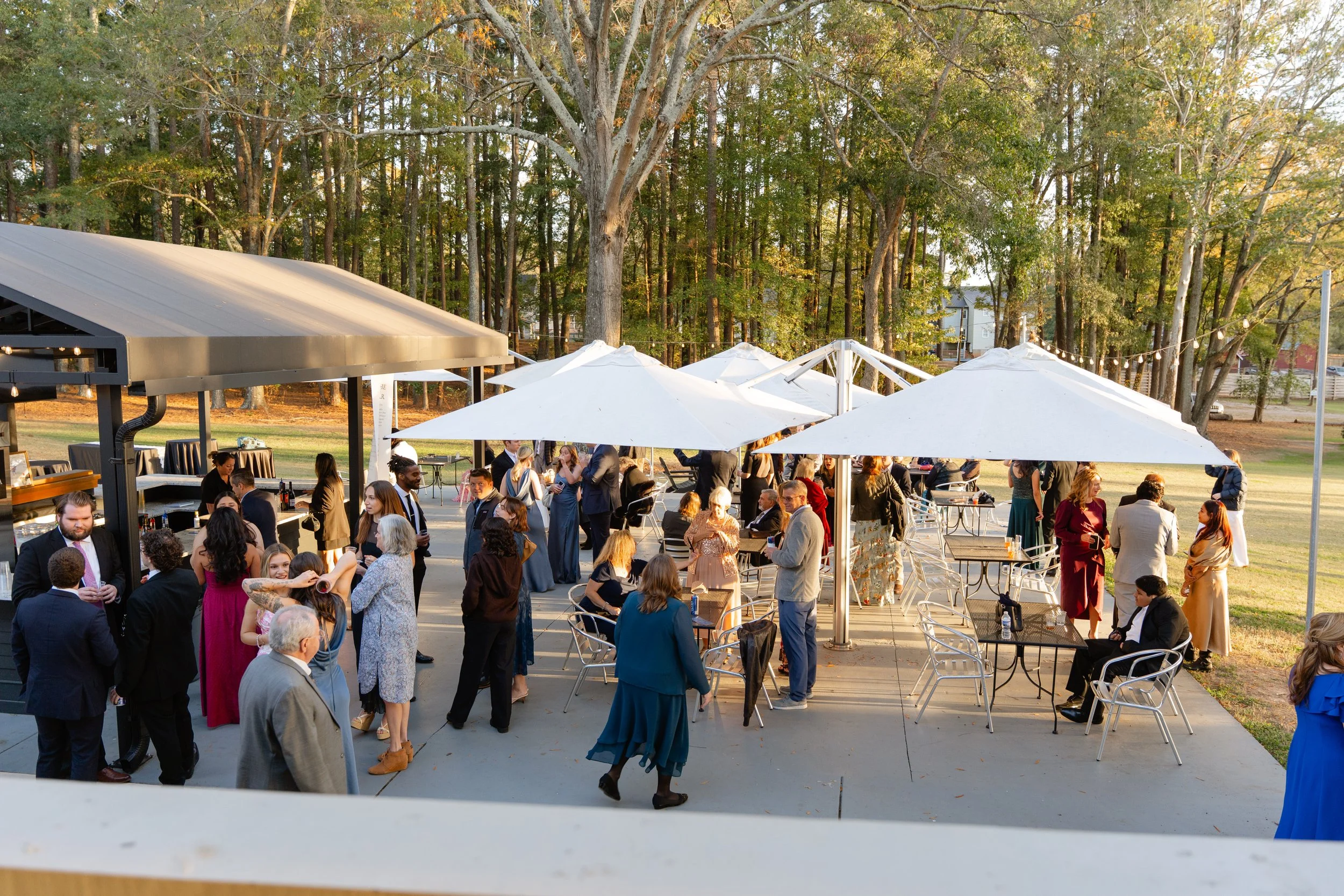 People gathered outdoors at a social event or party, with some standing and some sitting under white umbrellas, in front of trees, with a building and string lights in the background.