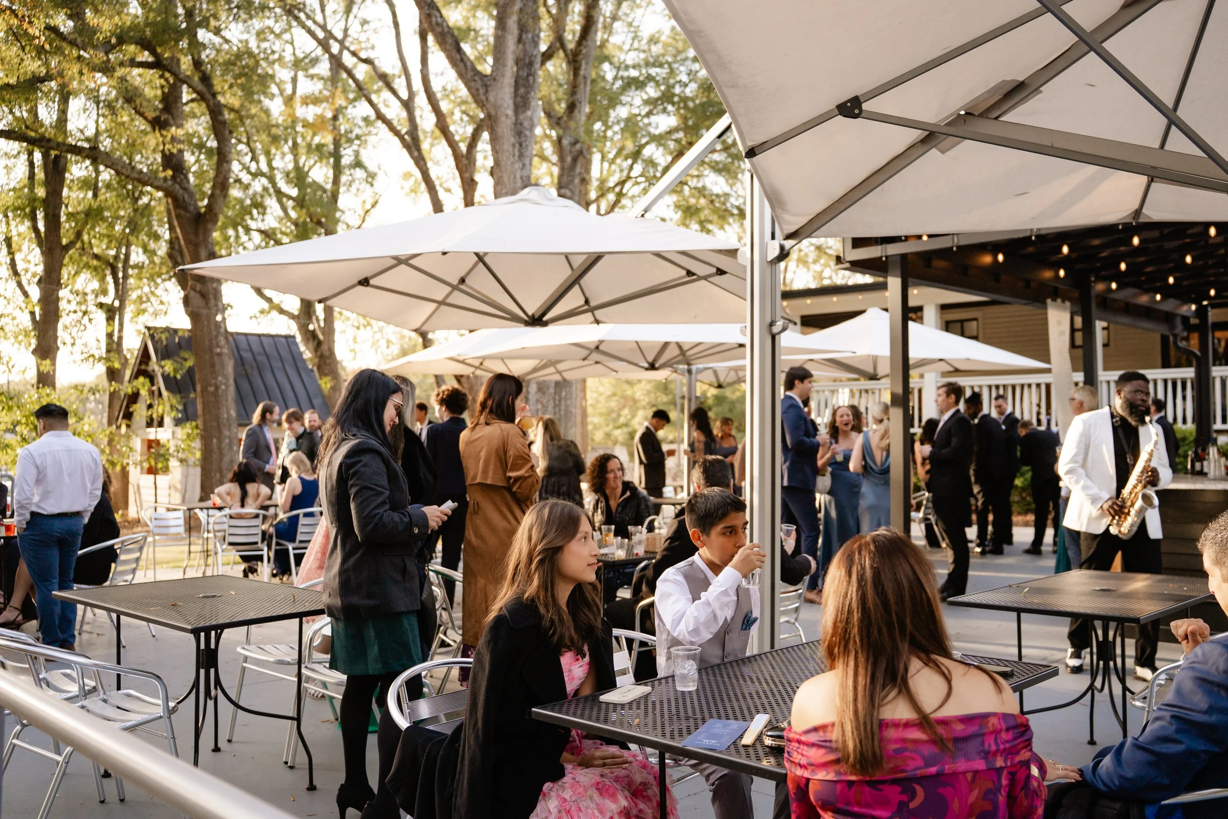 People gathered at an outdoor party or event under large white umbrellas and string lights, with some seated at tables and others standing or walking around, enjoying food and music.