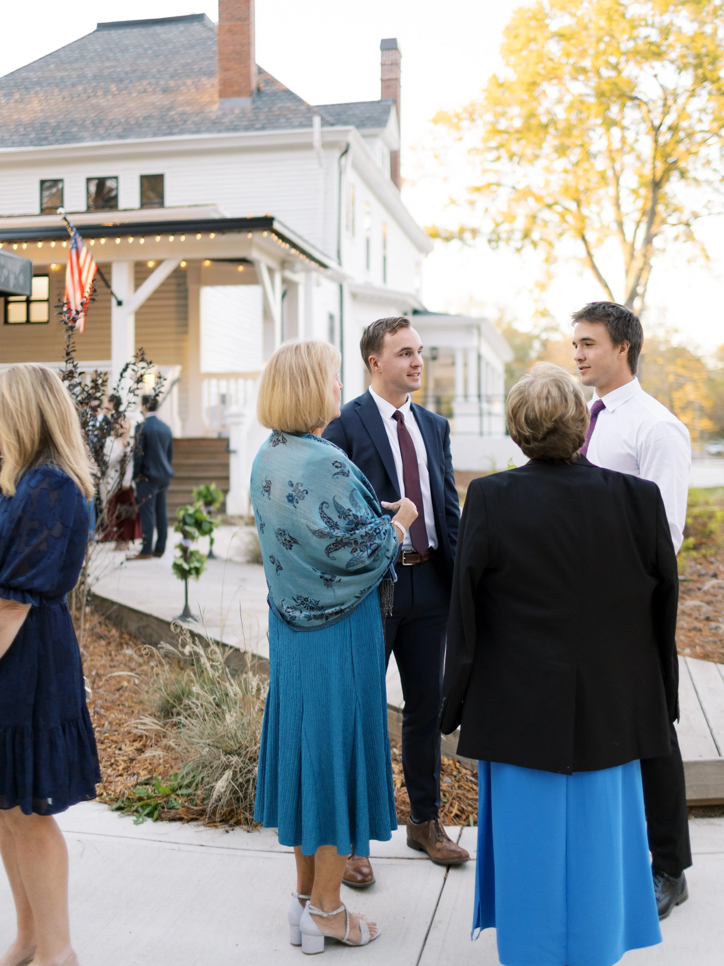 A group of people talking outside a white house with steps and string lights, during fall with trees showing autumn leaves.