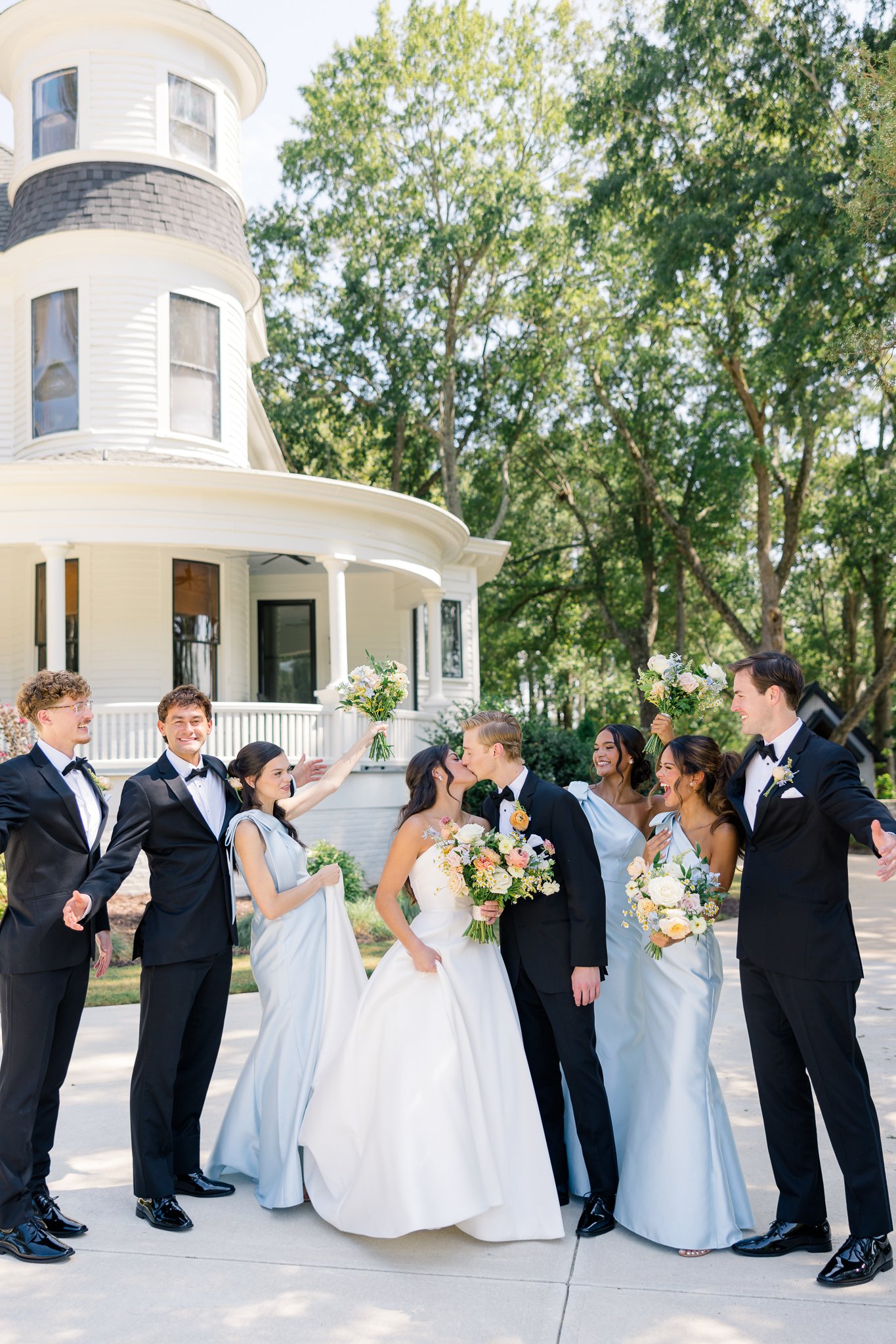 A wedding party outside a large white house with tall trees in the background, featuring a bride and groom kissing surrounded by bridesmaids and groomsmen celebrating.