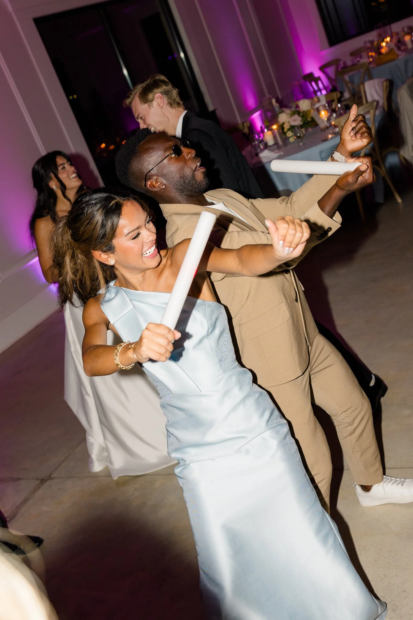 People dancing at a decorated party, holding white glow sticks, smiling and enjoying the celebration, with purple lighting and table settings in the background.