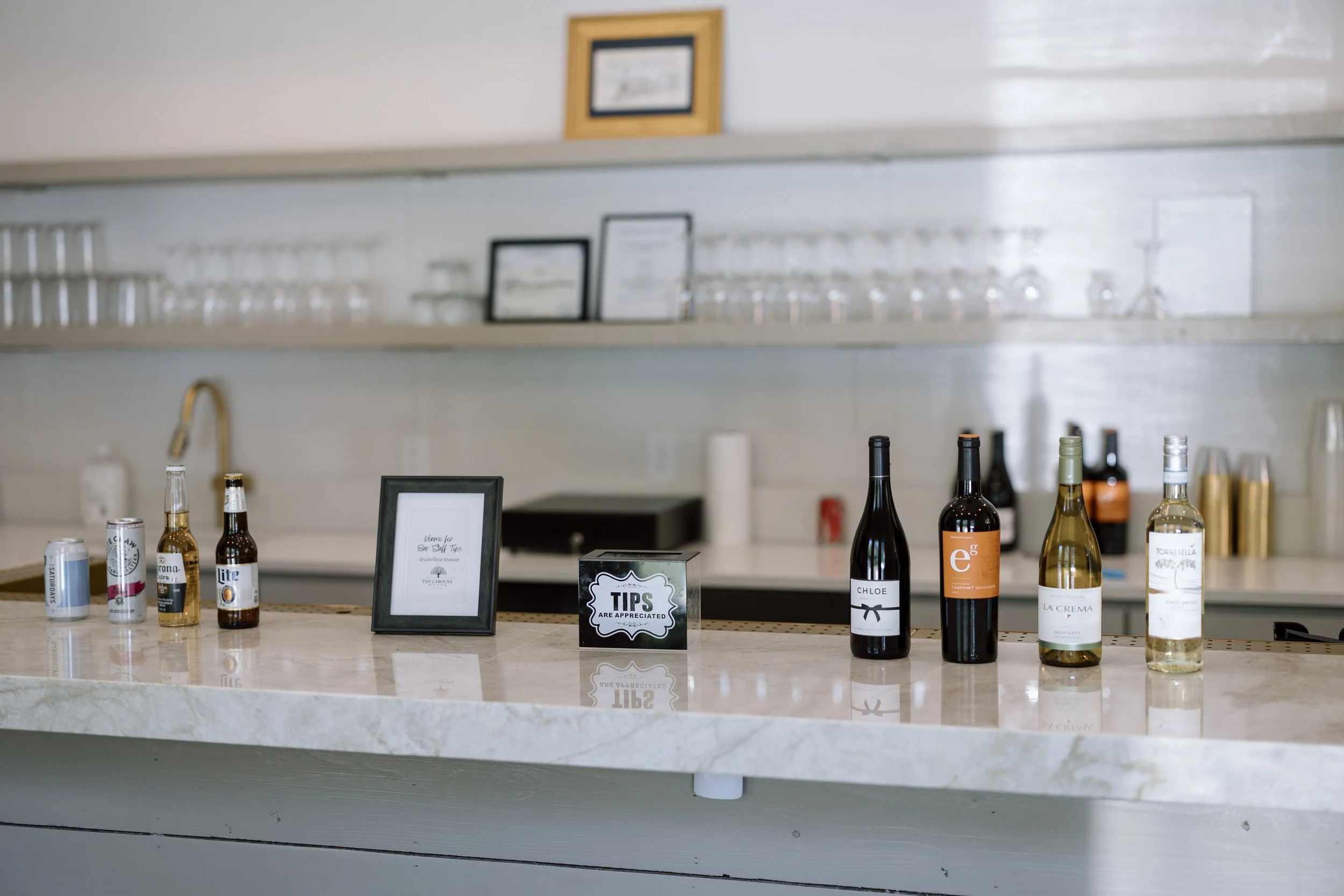 Bar counter with various bottles of drinks, a framed note, and a small sign that says 'Tips are appreciated', with glasses and awards on shelves in the background.