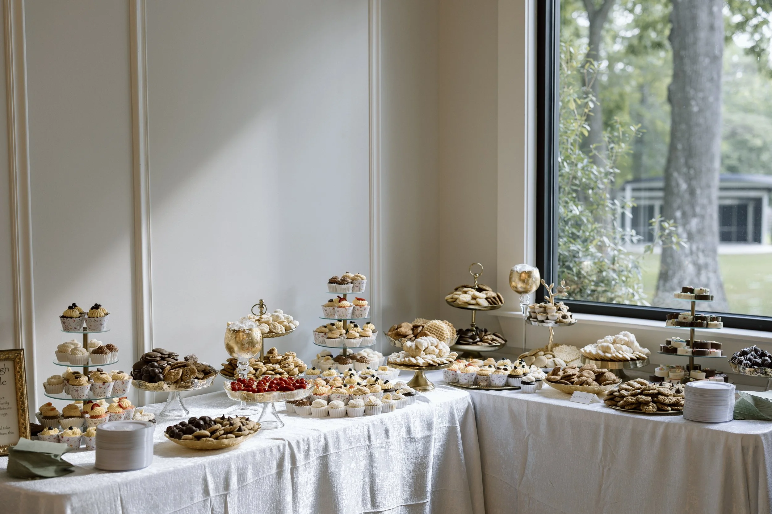 A dessert table with a variety of cookies, cupcakes, and chocolates set up near a large window with a view of trees outside.