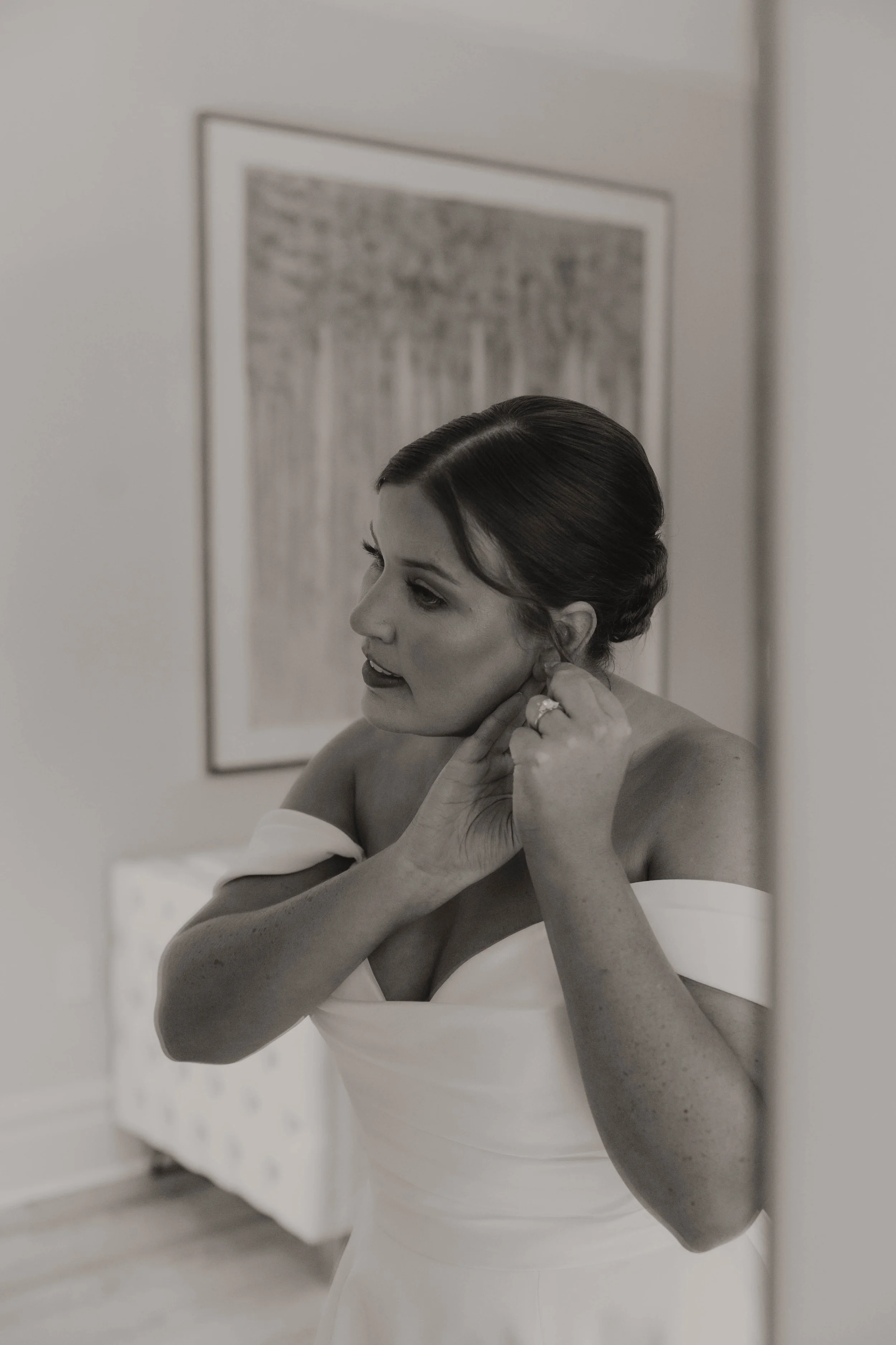 A woman in a wedding dress putting on an earring in front of a mirror.