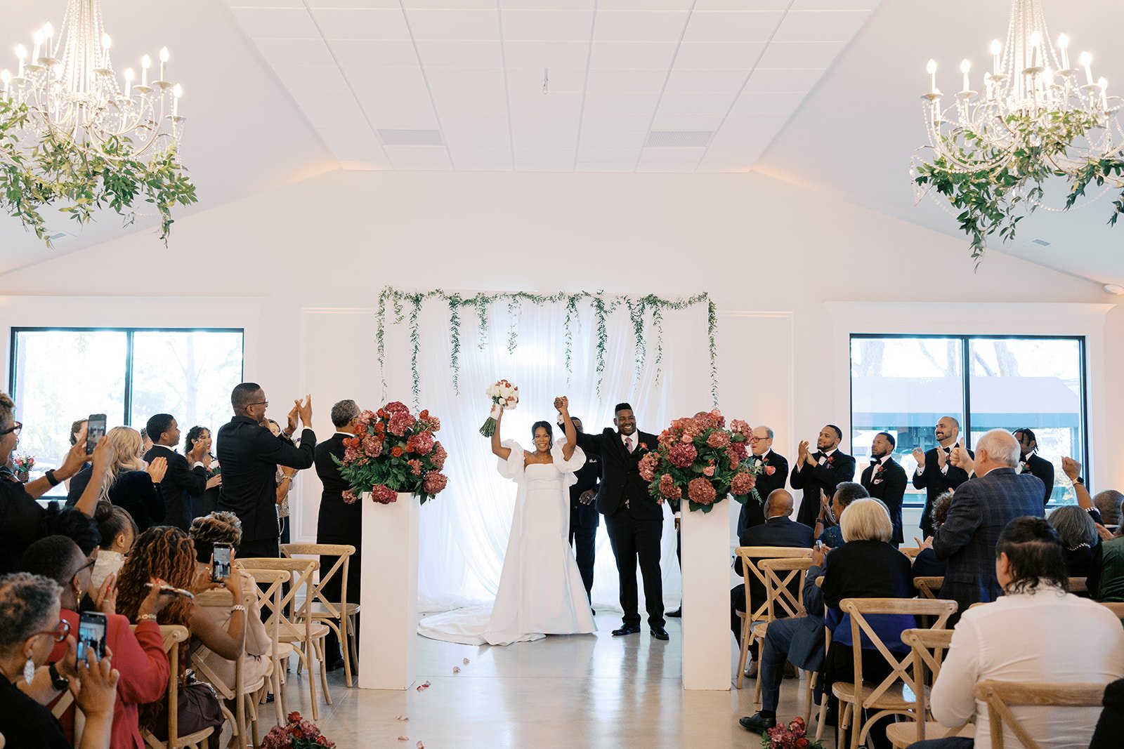 A wedding ceremony with a bride and groom standing in front of an audience, celebrating and raising their fists. The venue is decorated with large floral arrangements and greenery, with chandeliers hanging from the ceiling.