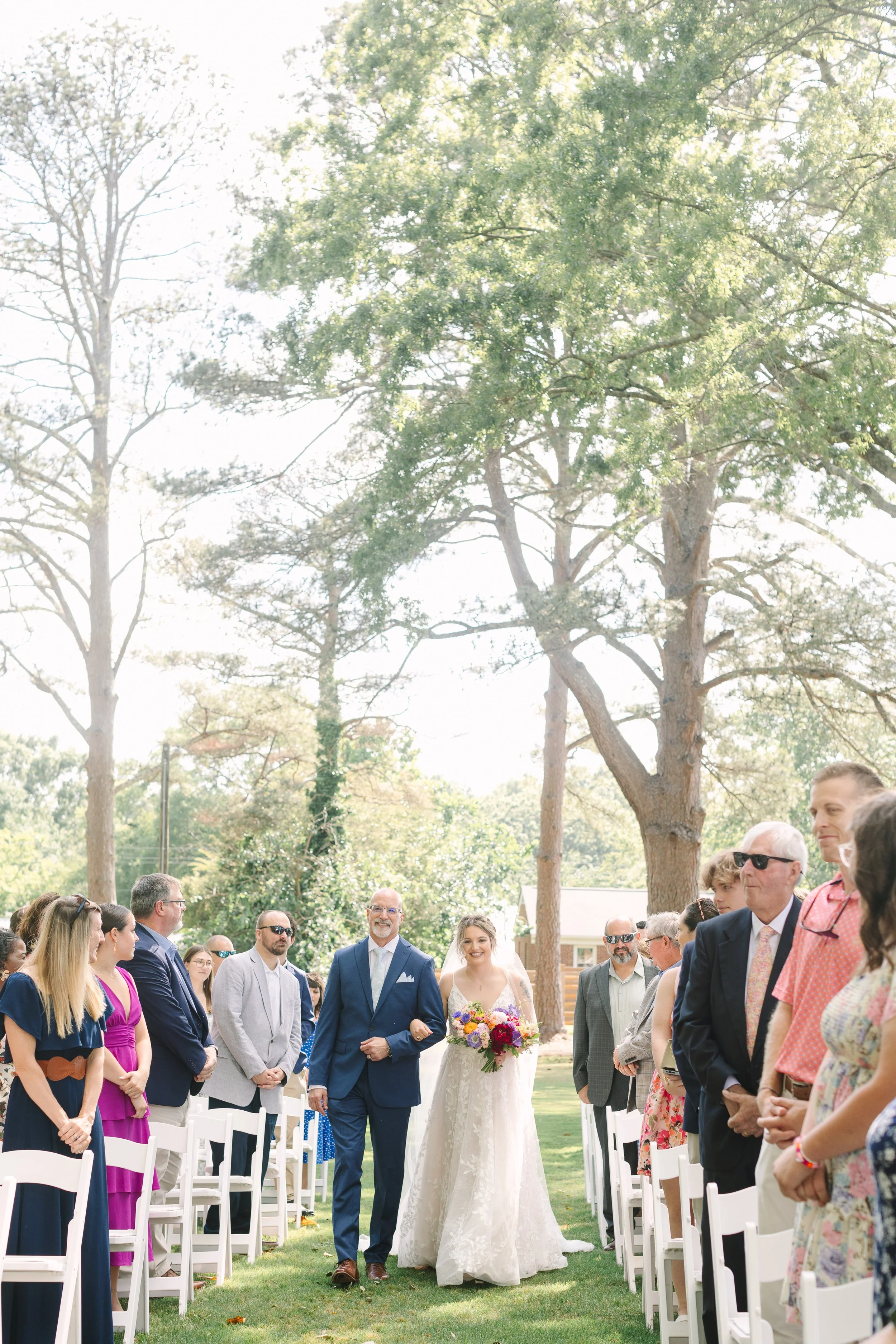 Bride walking down the aisle with her father at an outdoor wedding ceremony, surrounded by seated guests on a green lawn with tall trees in the background.