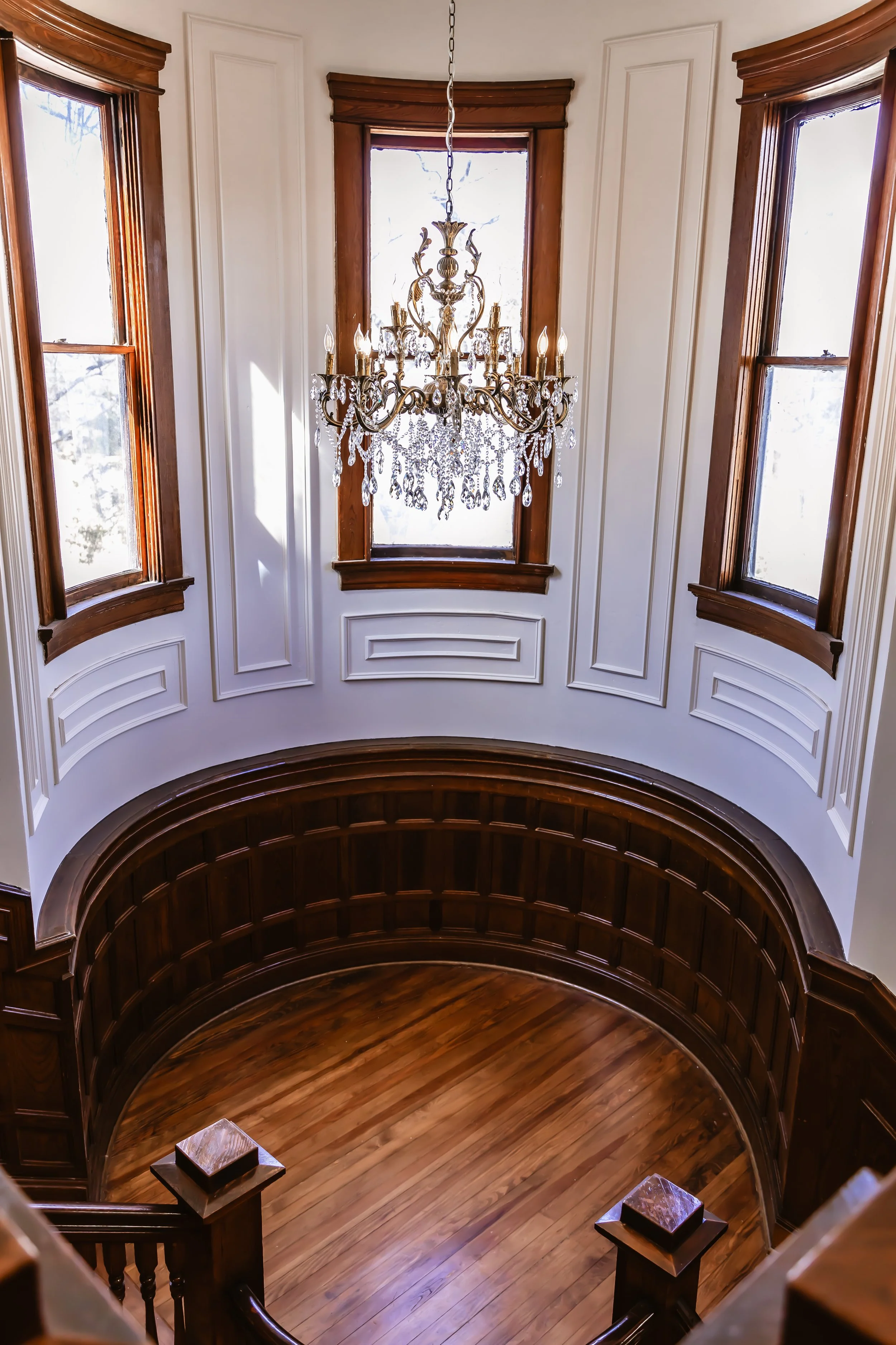 Interior view of a staircase area with a chandelier hanging from the ceiling, three large windows with wooden frames, and a curved wooden railing around the stairwell, also featuring white panel walls.