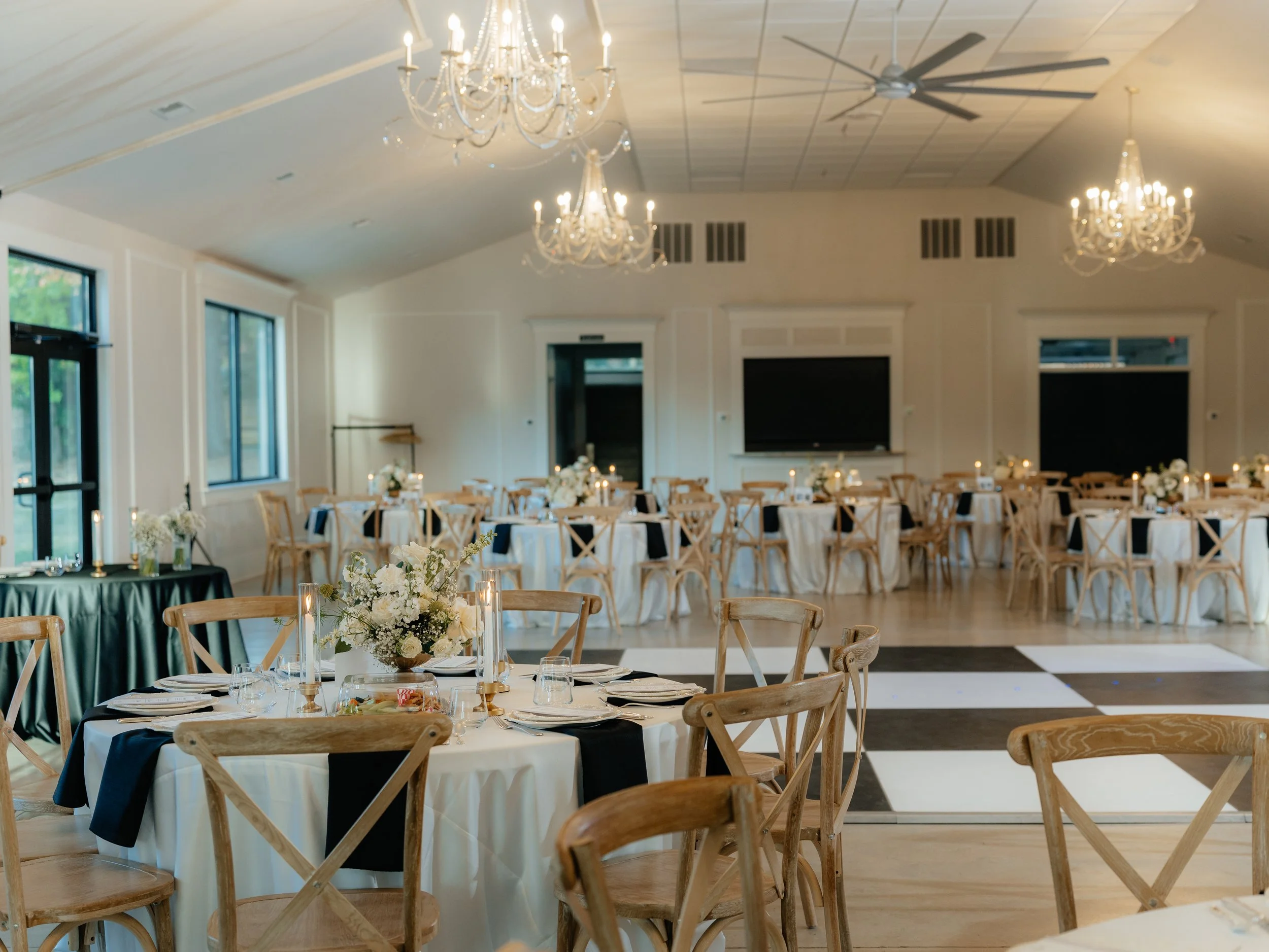 Elegant banquet hall decorated with chandeliers, large windows, and round tables covered with white tablecloths and navy napkins, adorned with floral centerpieces and candles, ready for an event.