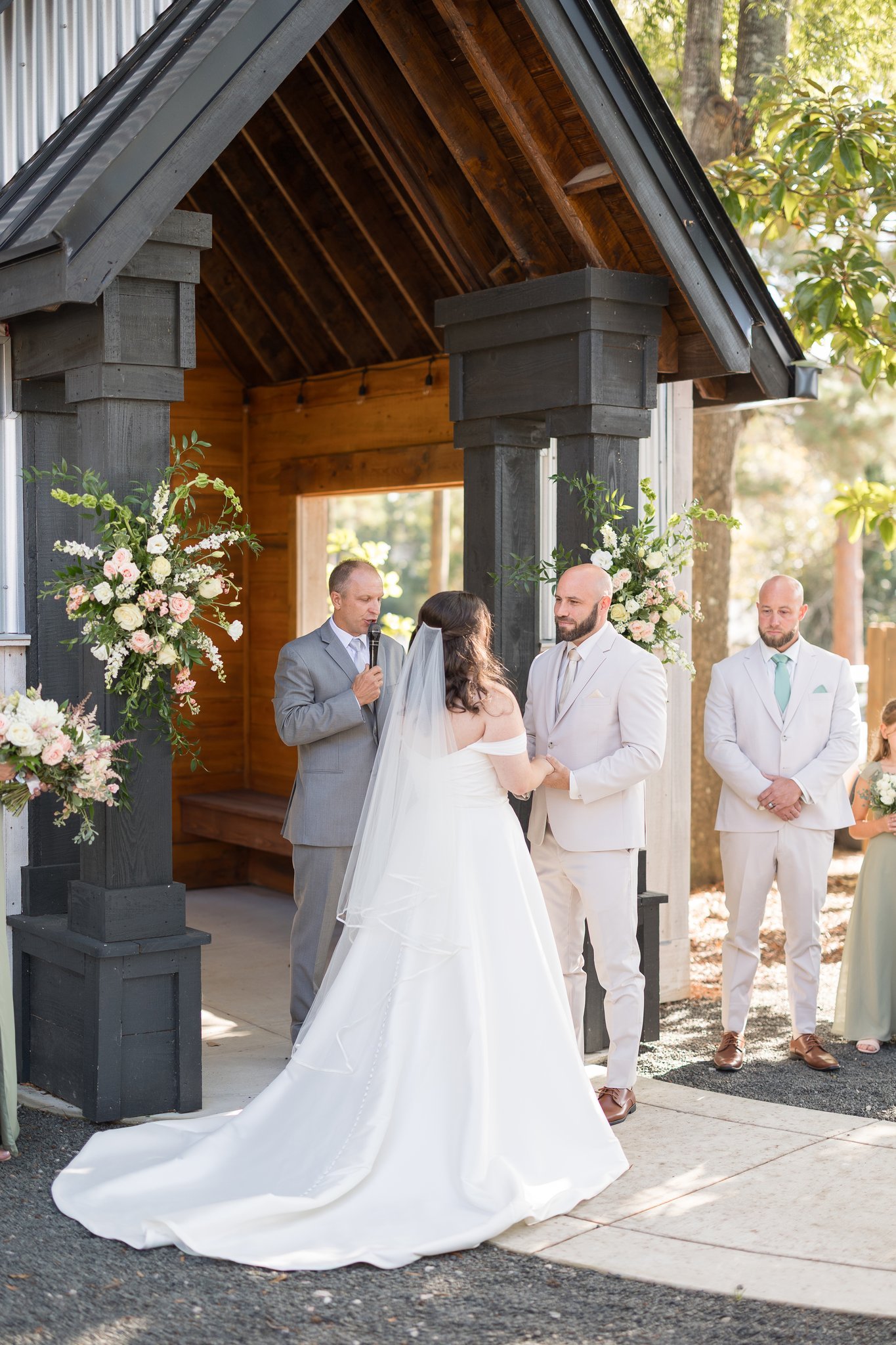 A couple getting married in an outdoor ceremony, with the bride wearing a white wedding gown and veil, and the groom in a white suit, holding hands in front of a officiant during the vows. Bridesmaids and groomsmen are standing nearby, and the ceremo