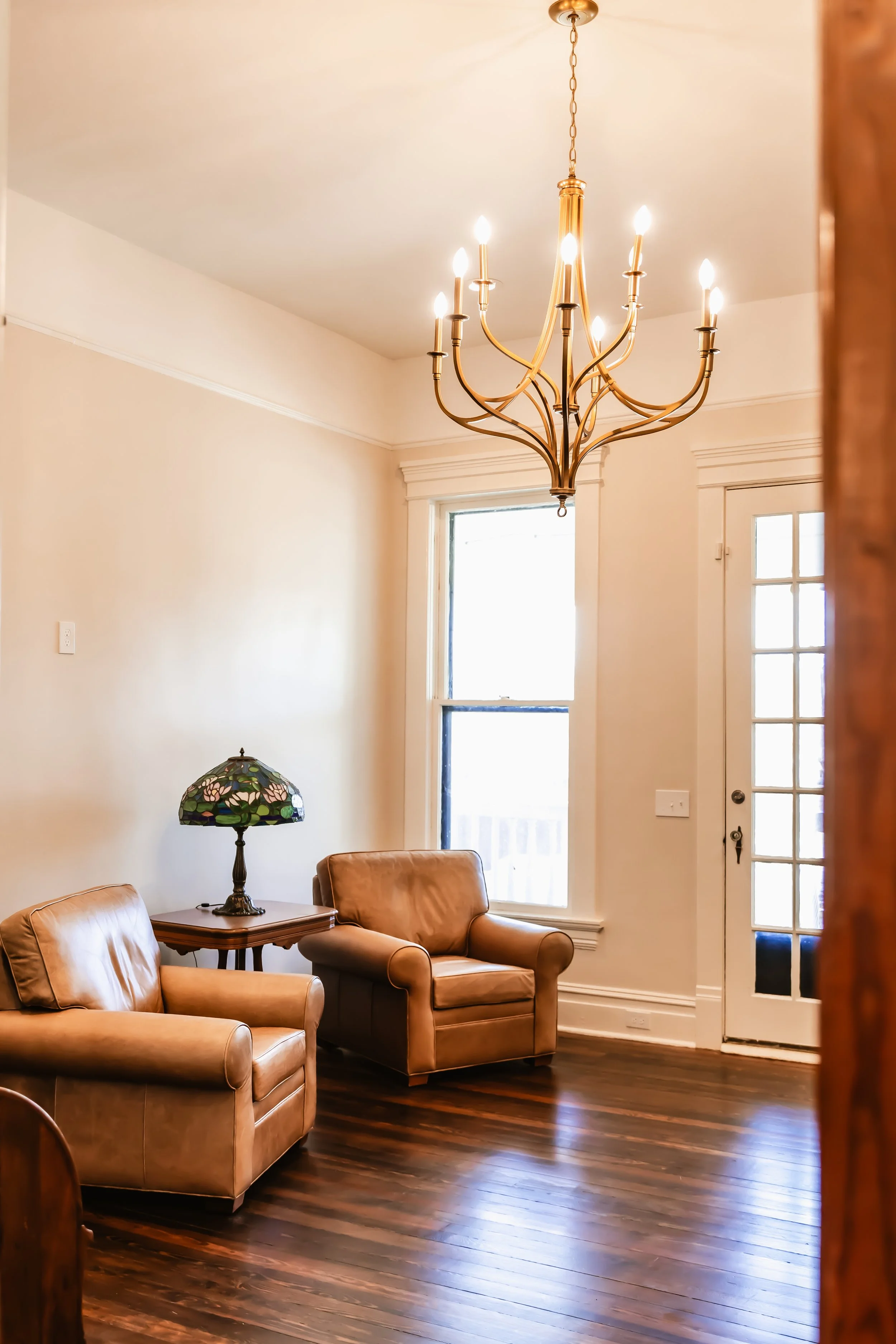 Living room with two tan leather armchairs, wooden floor, a side table with a stained glass lamp, a large window, a glass-pane door, and a gold chandelier hanging from the ceiling.
