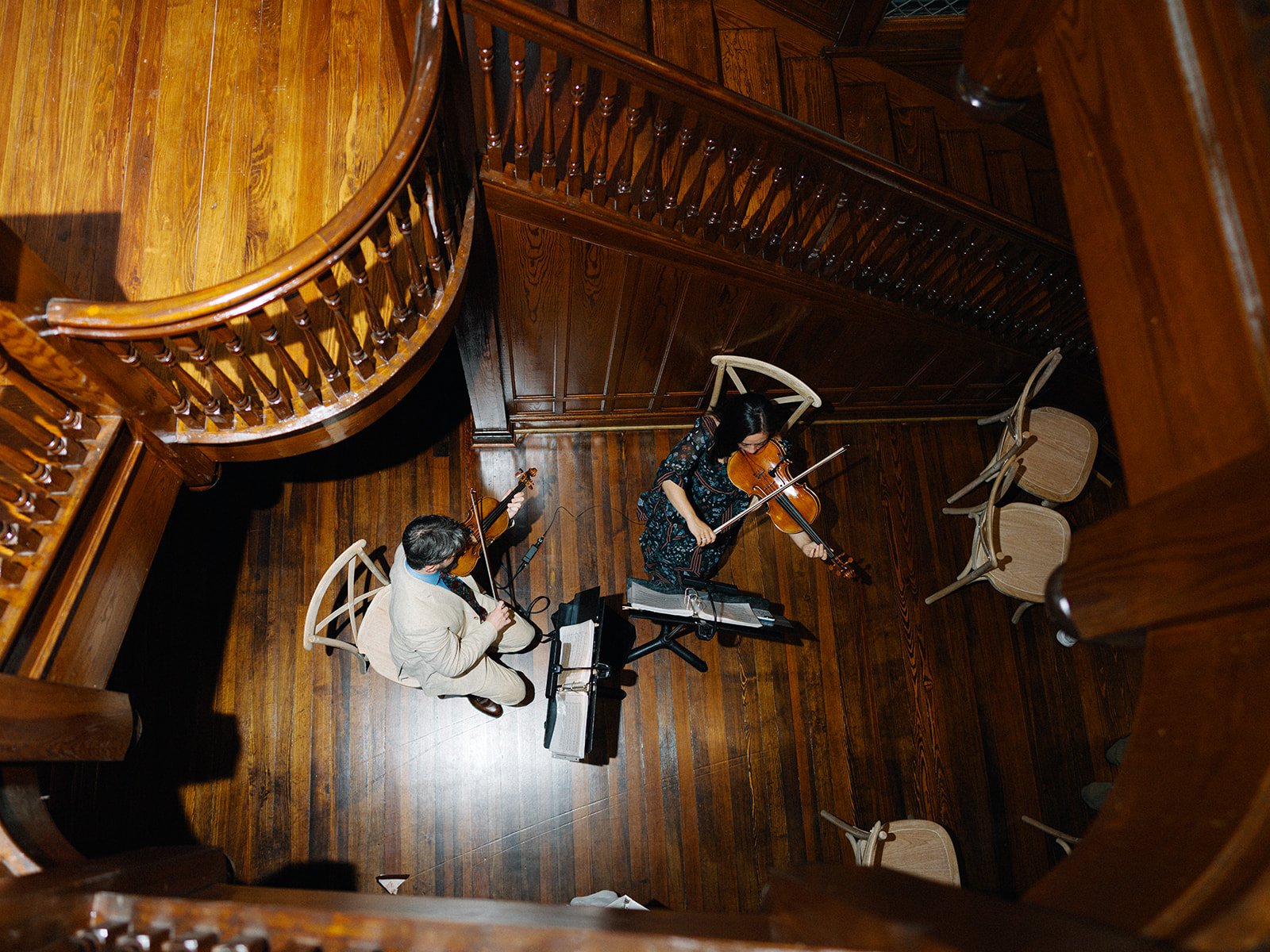 Two musicians playing violins in a wooden-floored, intimate concert space with wooden chairs and a wrap-around balcony.