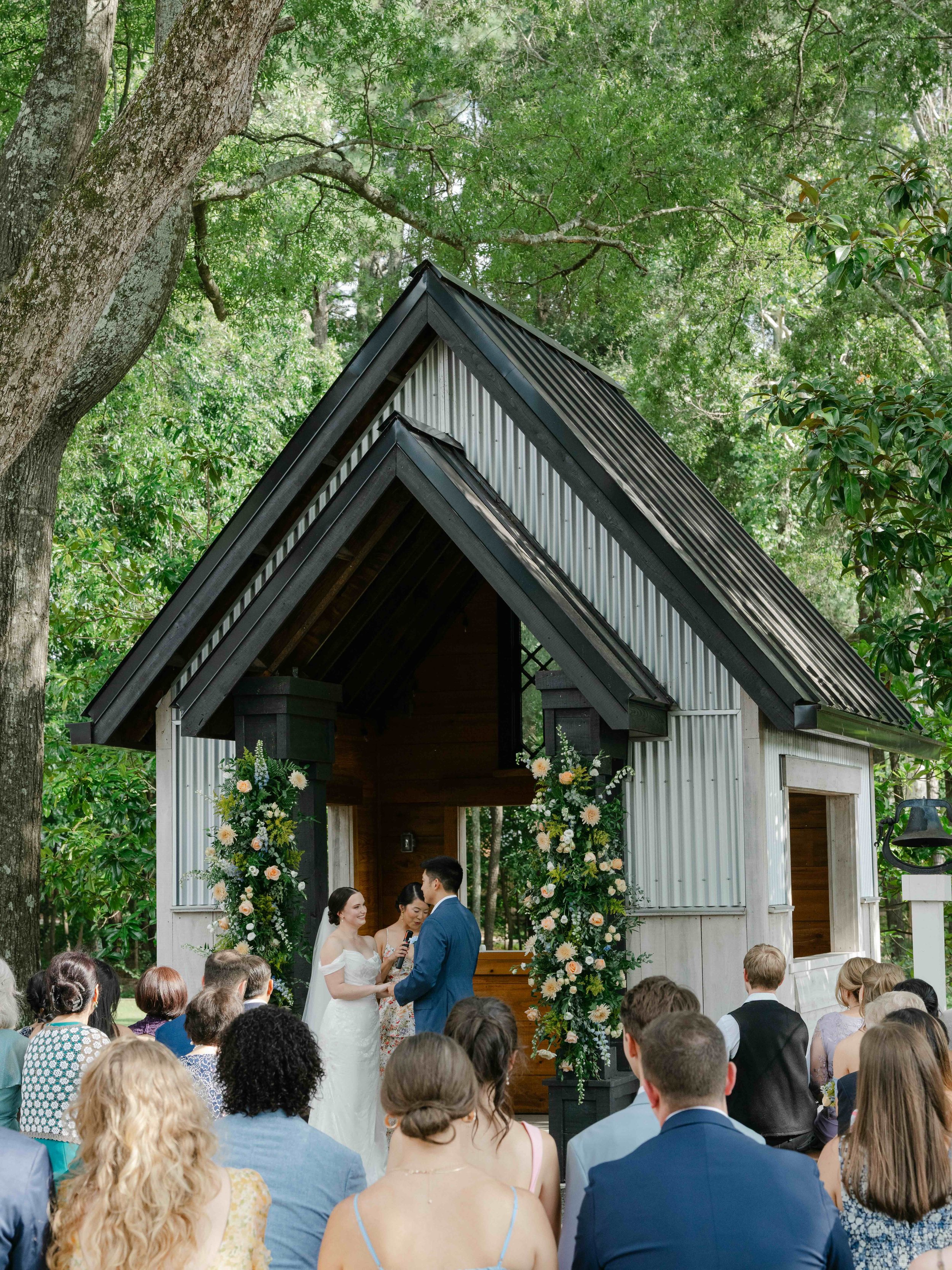 A wedding ceremony outdoors with a bride and groom exchanging vows under a small wooden chapel decorated with flowers, surrounded by green trees and seated guests.