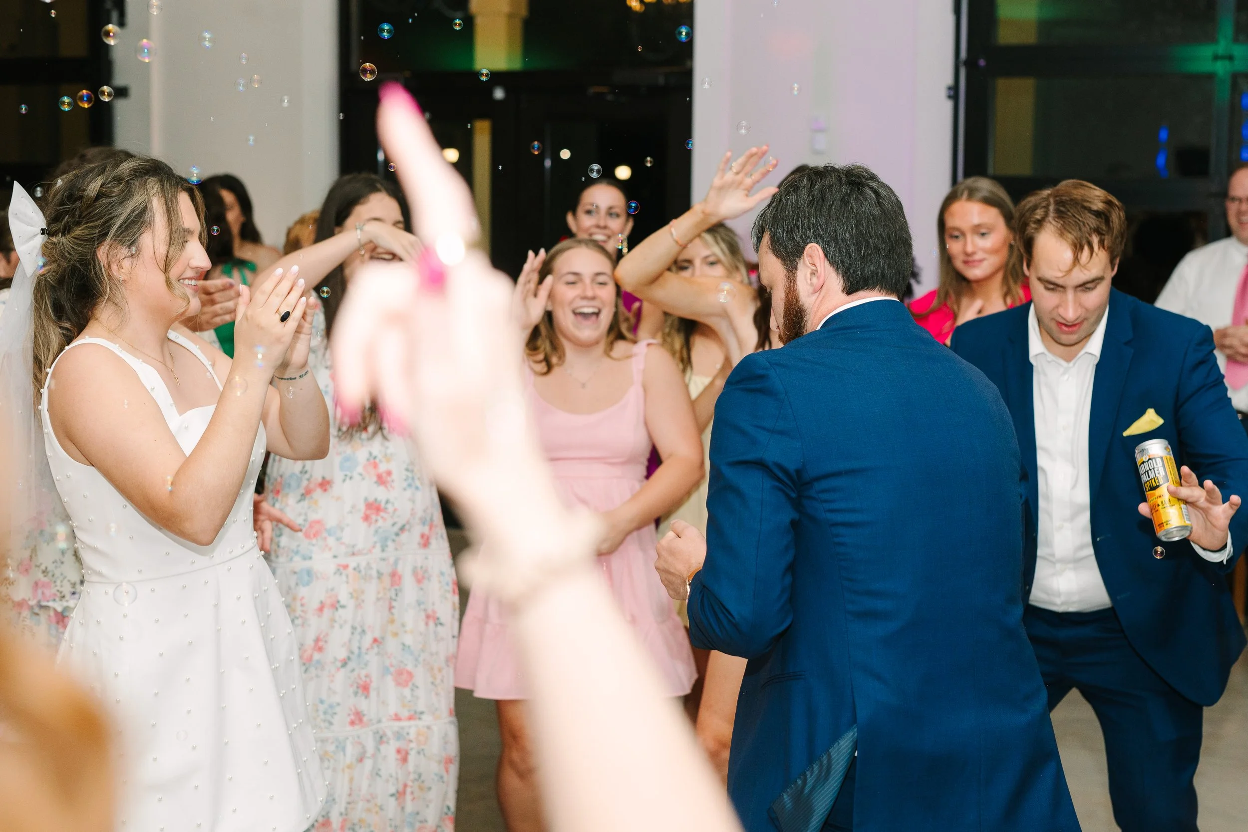 People celebrating at a party, with two men in suits and women in colorful dresses, surrounded by bubbles.