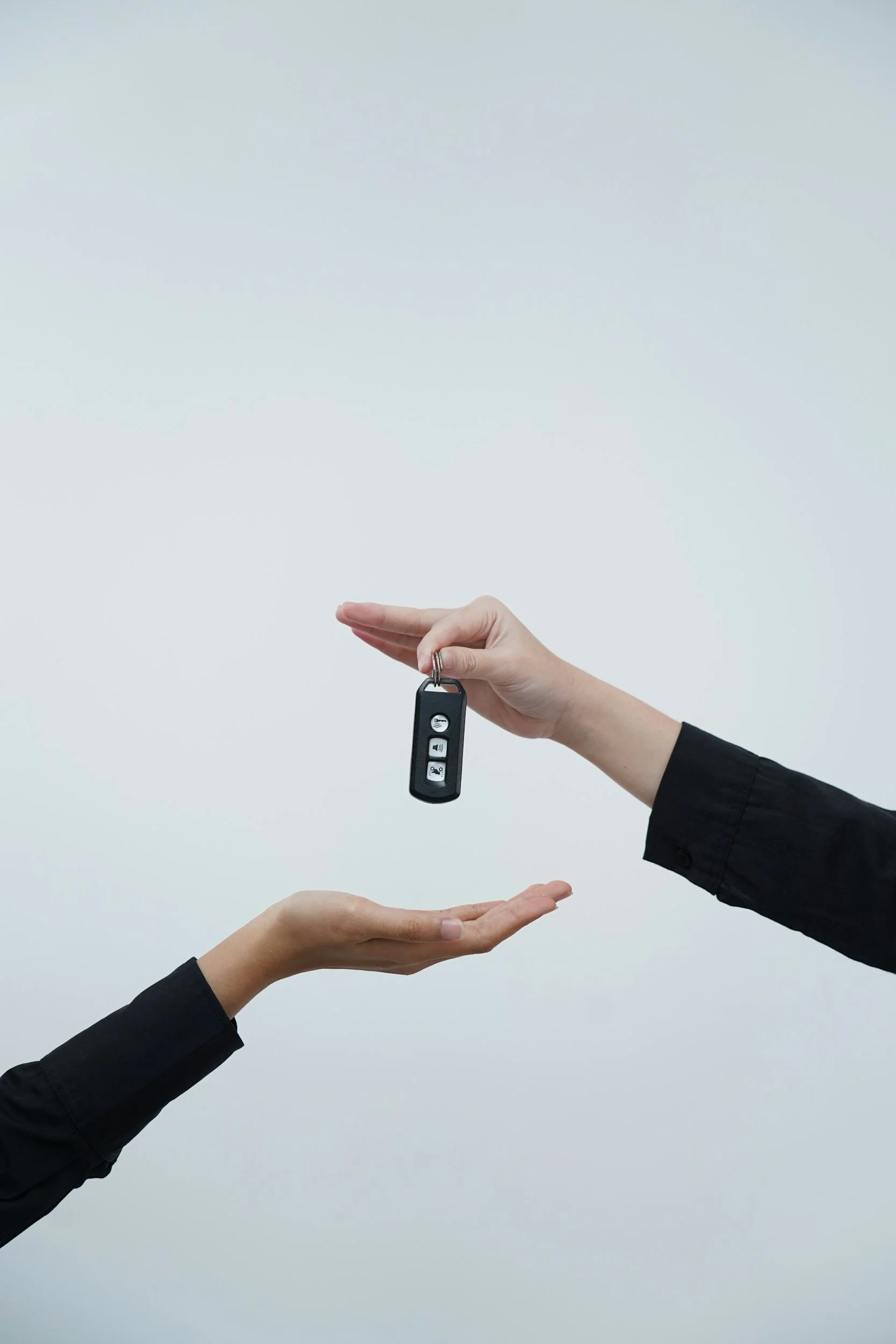 A person in a black shirt hands a key fob with three buttons to another person, who has hands extended to receive it, against a plain light blue background.