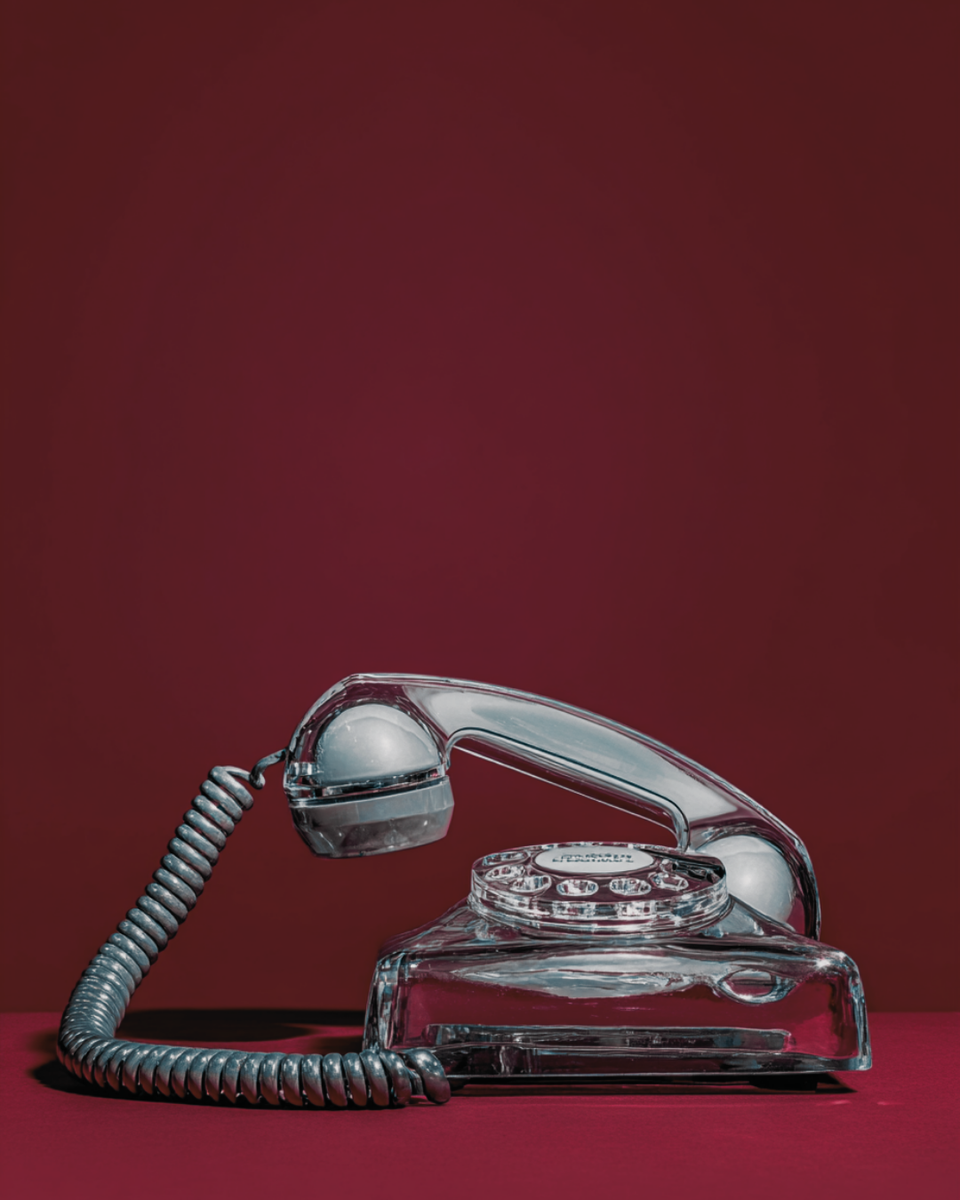 Vintage chrome rotary telephone with a transparent glass body against a deep burgundy background — editorial brand image for It's Cindy Kumar