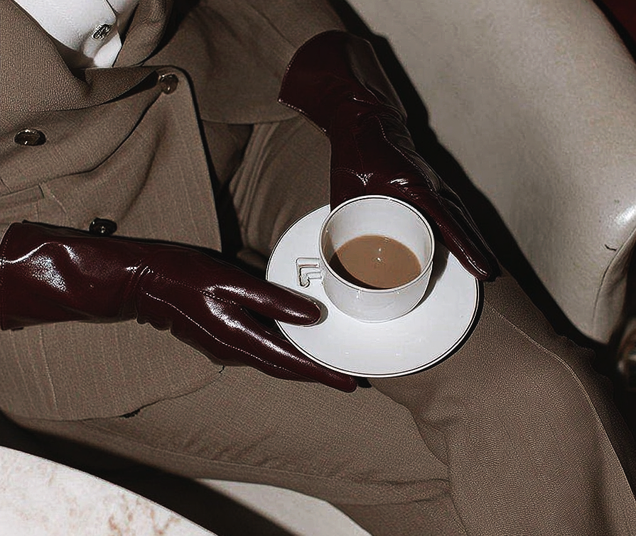 Dark burgundy leather-gloved hand reaching toward a white fine china teacup and saucer on a neutral surface — editorial brand image for It's Cindy Kumar, growth advisory and profit strategy