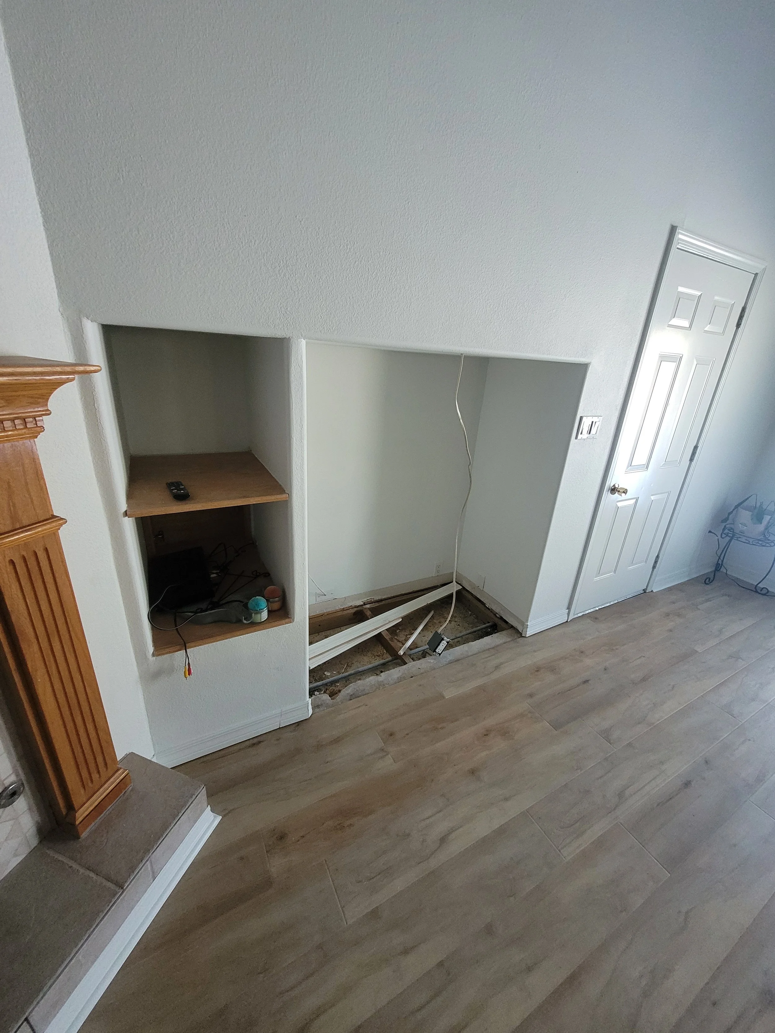 Room under renovation with an open space in the wall, exposing electrical wiring and plumbing. A white door is on the right, and the floor is covered with light wood planks.