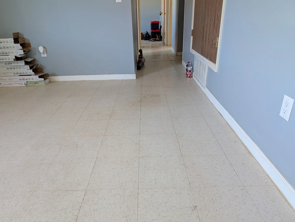 Empty room with beige tiled floor, light blue walls, and some boxes and tools near the back wall.