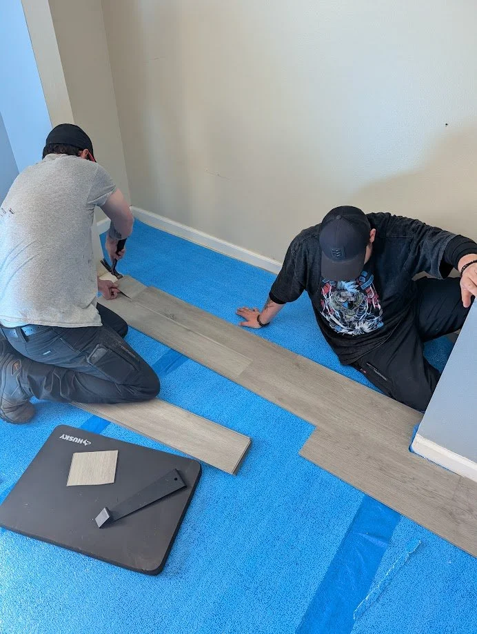 Two people installing a wood or vinyl flooring strip in a room with blue protective tape on the floor.