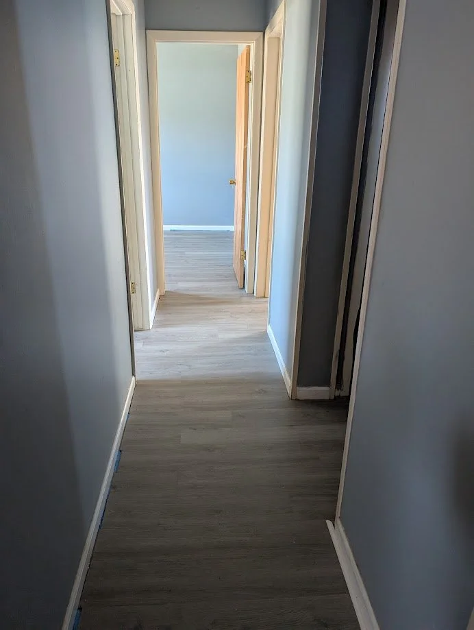 A narrow hallway in a house with light blue walls, white trim, and wood-look flooring, leading to an open door at the end.