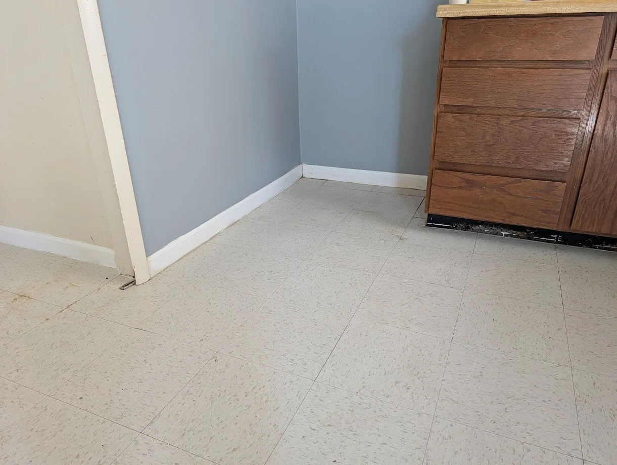 Corner of a room with beige and blue painted walls, white baseboards, and a beige tiled floor. Part of a wooden cabinet is visible in the right side of the image.