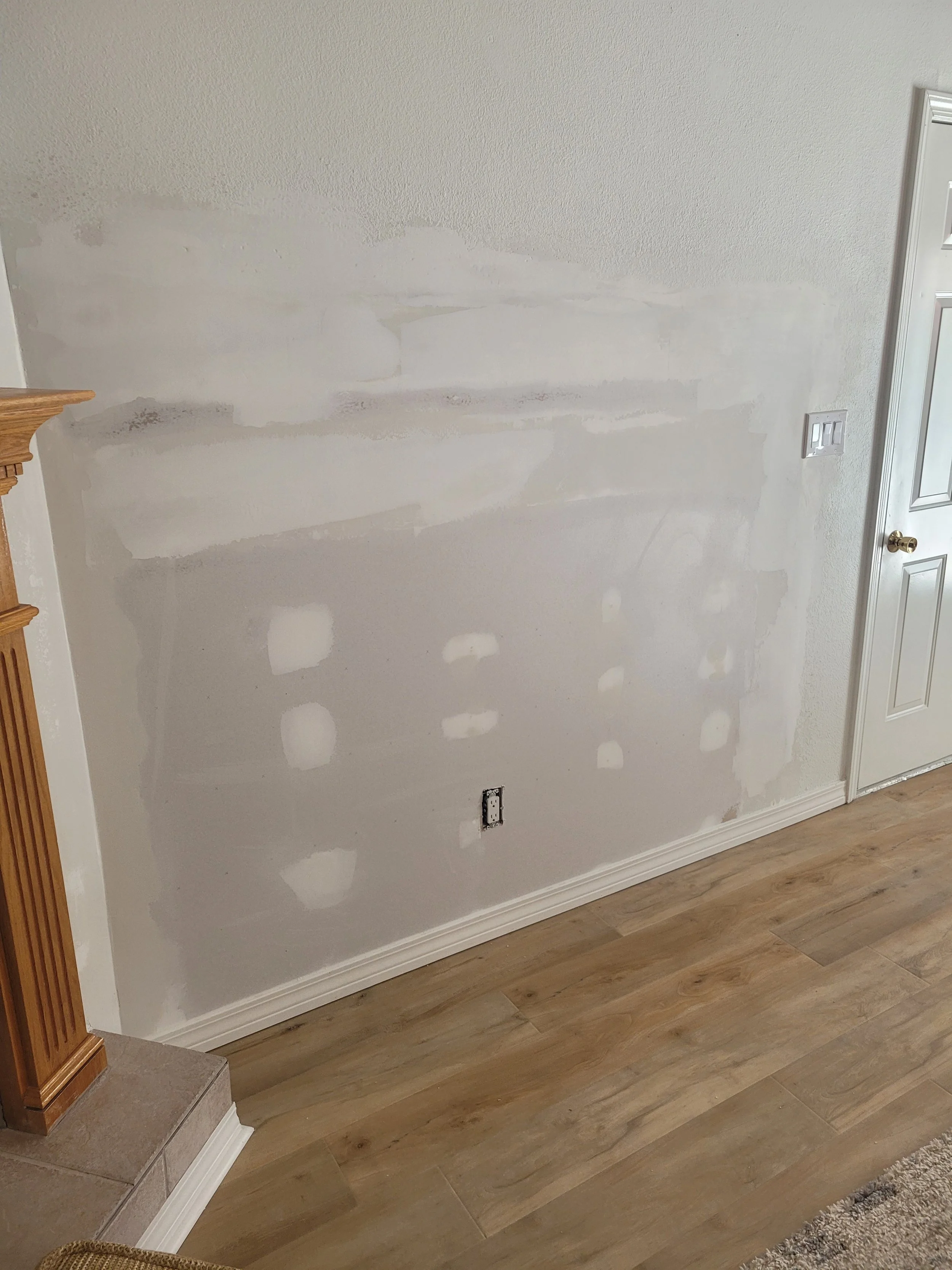 Living room wall with patchy drywall repairs, a partially painted section, and an electrical outlet without a cover. Part of a wooden staircase and beige carpet are visible.
