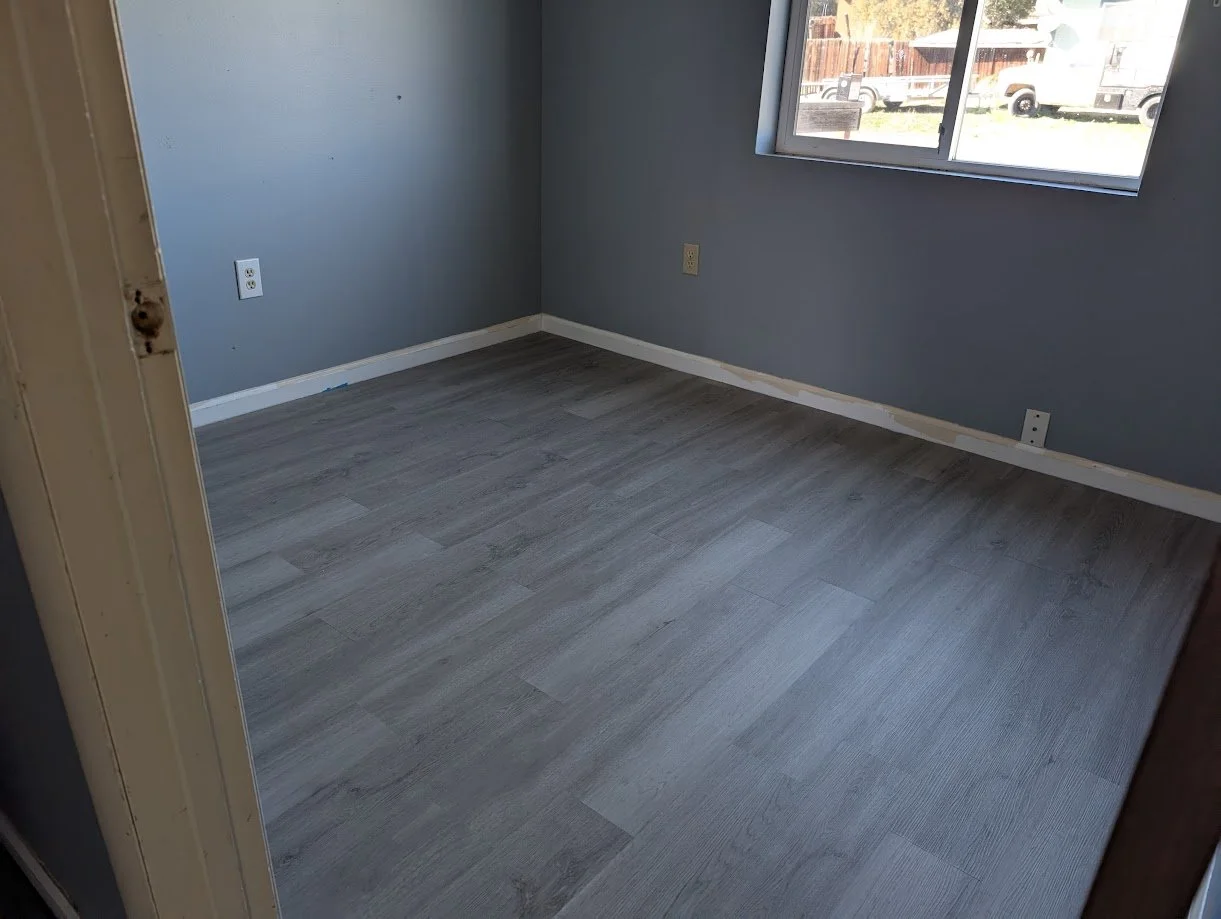 Empty room with blue walls, gray flooring, a large window, and white baseboards.