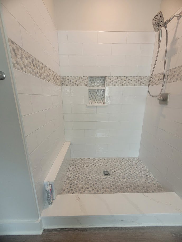 Empty walk-in shower with white tiles, decorative gray strip, small mosaic tiles on the floor and in a niche, and a handheld showerhead.