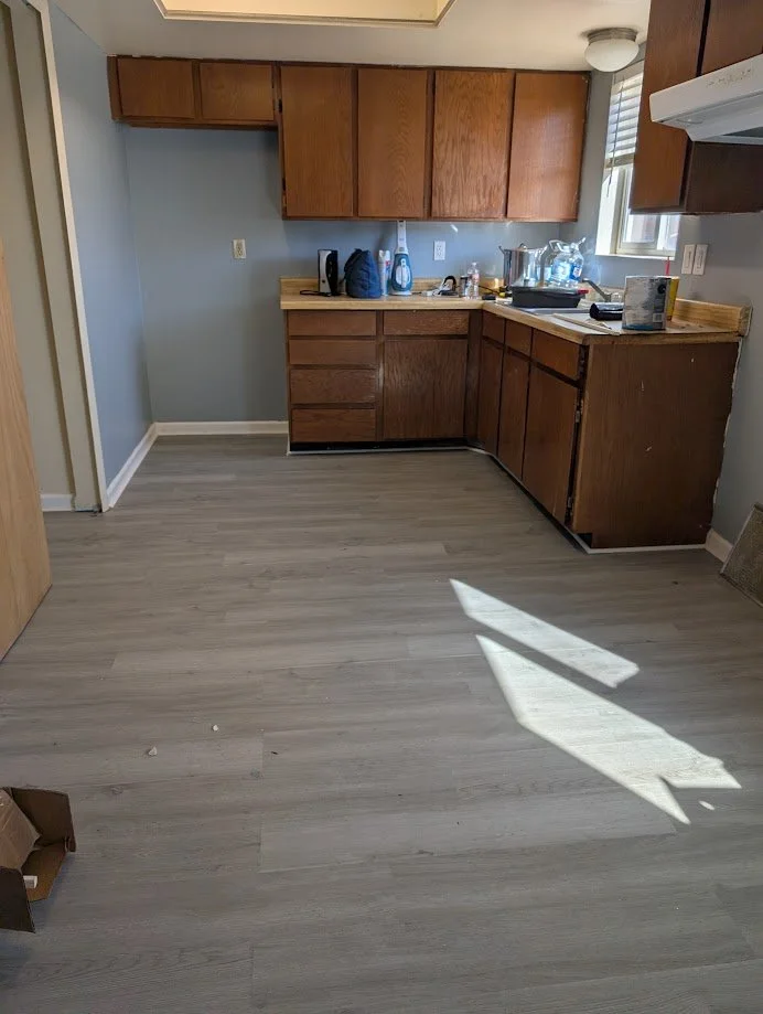 Empty kitchen with wooden cabinets, a countertop with appliances, and a window letting in sunlight on light-colored flooring.