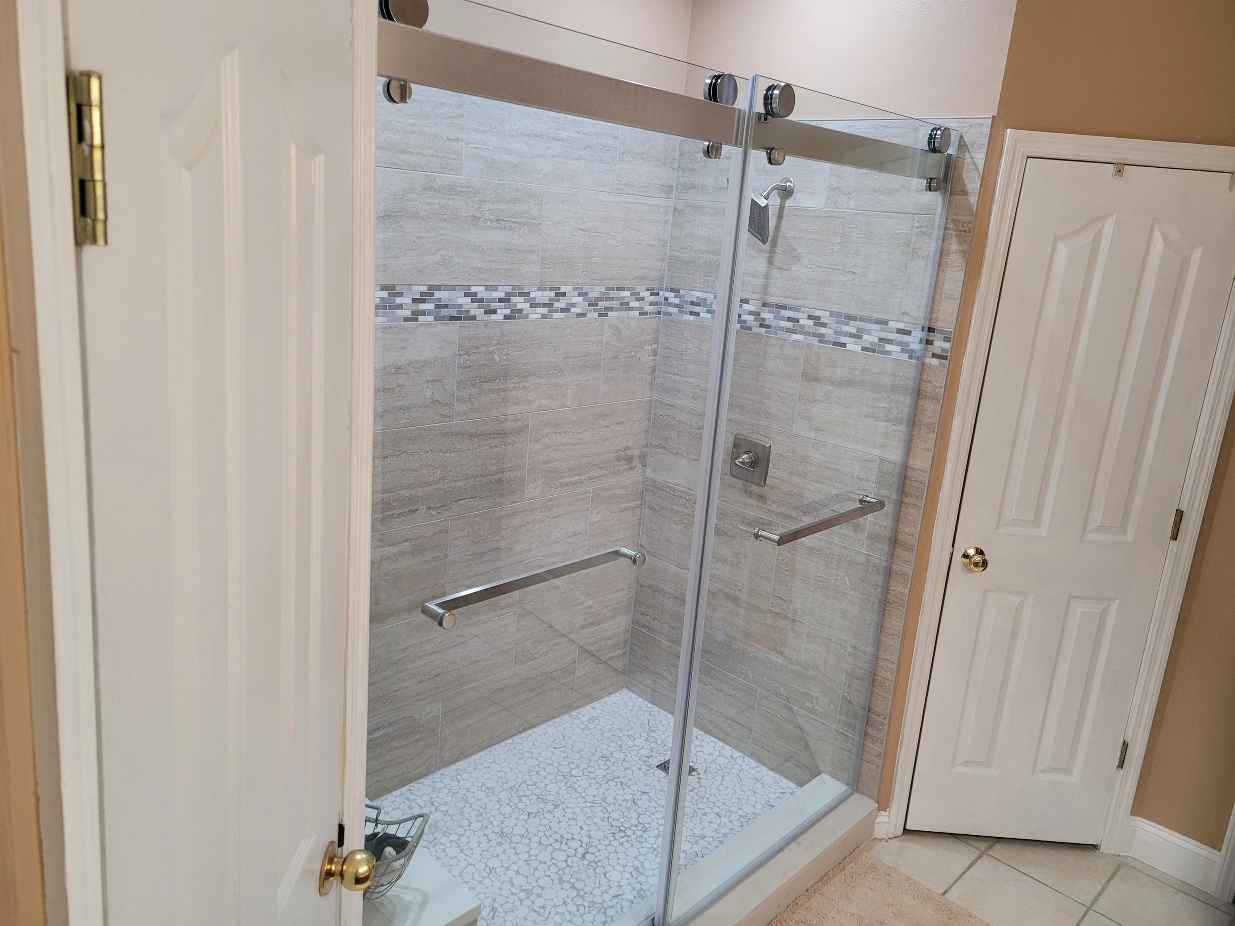 Bathroom shower with a glass door, beige tiled walls with a decorative mosaic strip, a square shower head, and a grab bar.