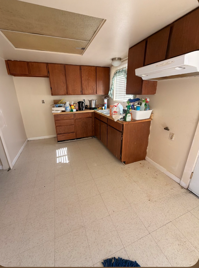 Empty kitchen with wood cabinets, cluttered countertop, and stained beige floor tiles.