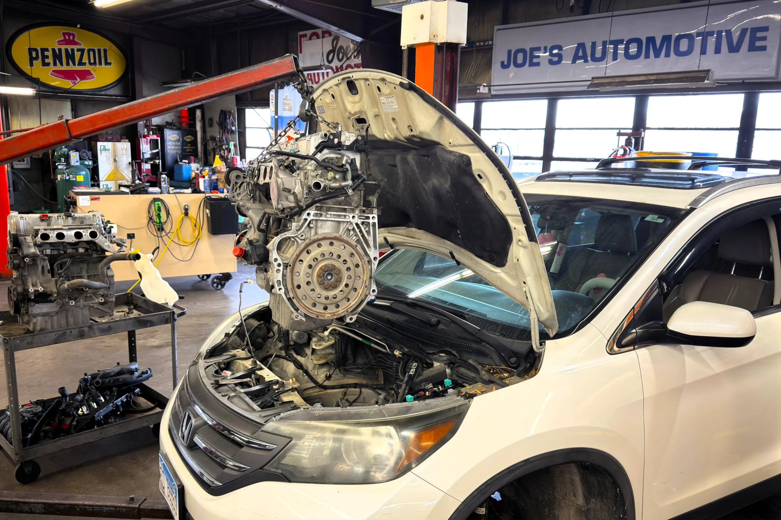 An automotive repair shop with a white Honda vehicle having its hood raised and engine removed, a crane hoist lifting the engine out, tools and engine parts nearby, and signs including Pennzoil and Joe's Automotive in the background.