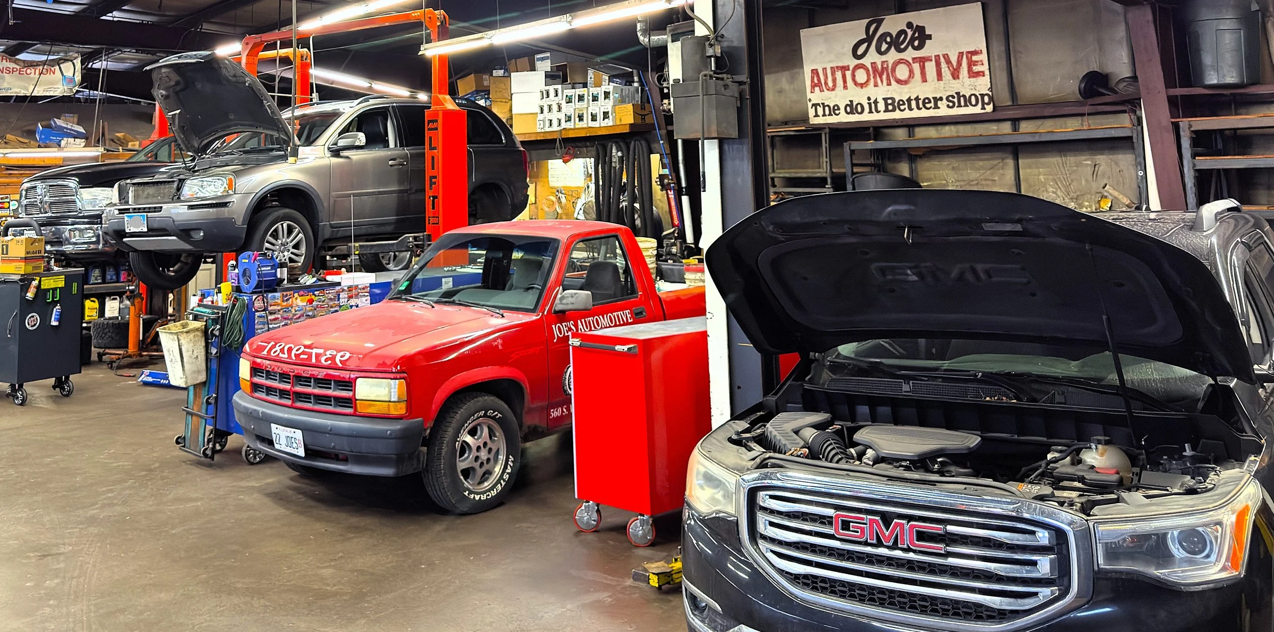 An auto repair shop with vehicles being serviced. A black GMC vehicle with an open hood in the foreground, a red pickup truck with Joe's Automotive signage, and a gray SUV on a lift in the background.