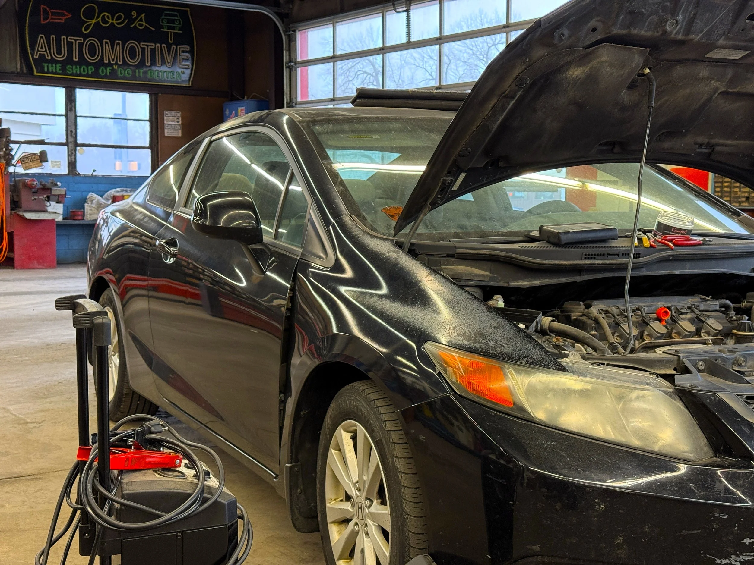 A black Honda car in a mechanic's garage with its hood open, undergoing maintenance. The garage has a sign that reads 'Joe's Automotive The Shop of 'Do It Better,' and various tools and equipment are visible.